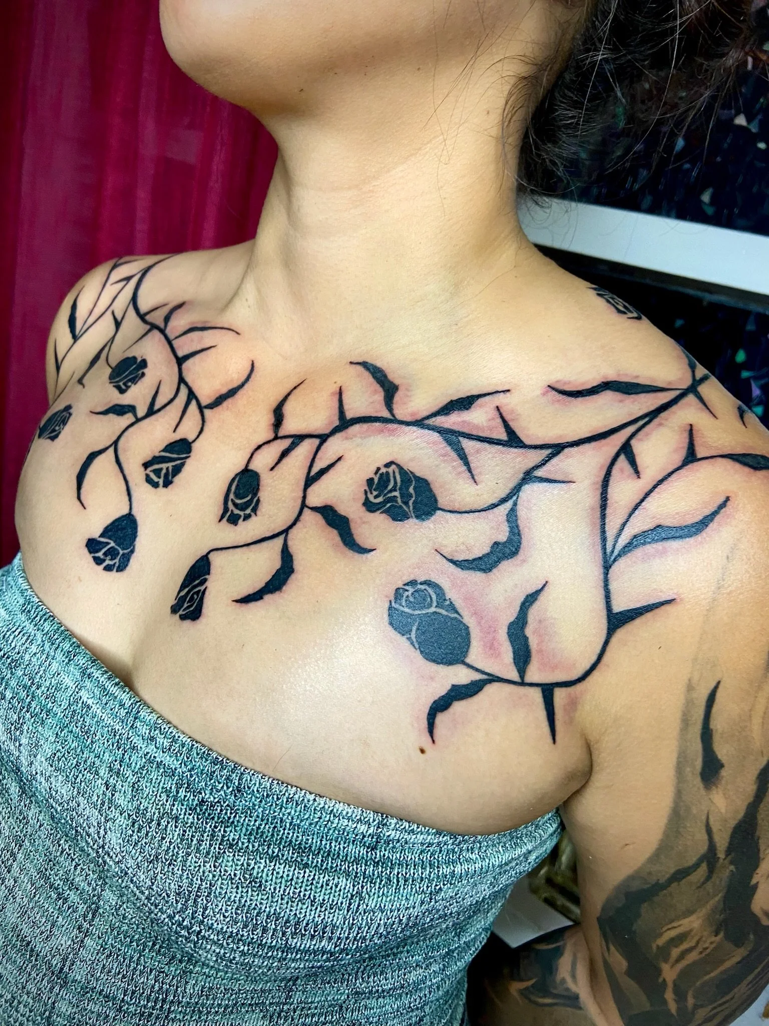 Close-up of a person's shoulder and collarbone area with a black floral tattoo, wearing a blue textured top, against a red background.