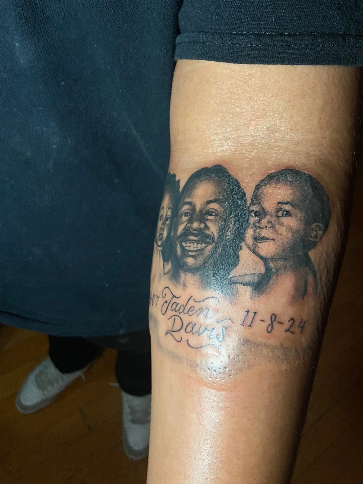 A tattoo on a person's forearm depicting three faces of women and a man, with a handwritten note below that reads 'In Loving Memory of Jaden, 11-8-24'.