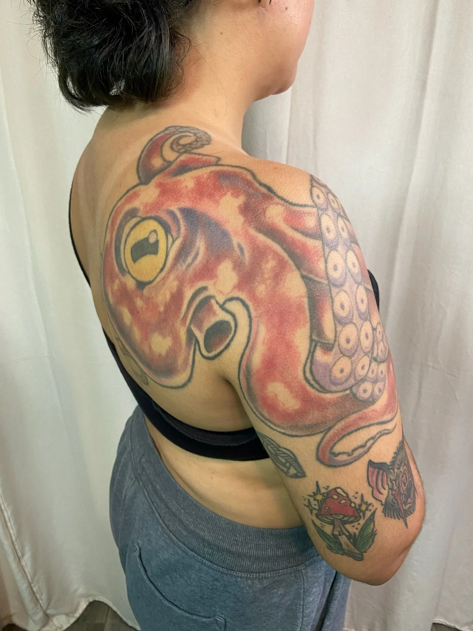 A person showing their left shoulder and upper arm with a large shoulder tattoo before the coverup, wearing a black bra strap and gray sweatpants.