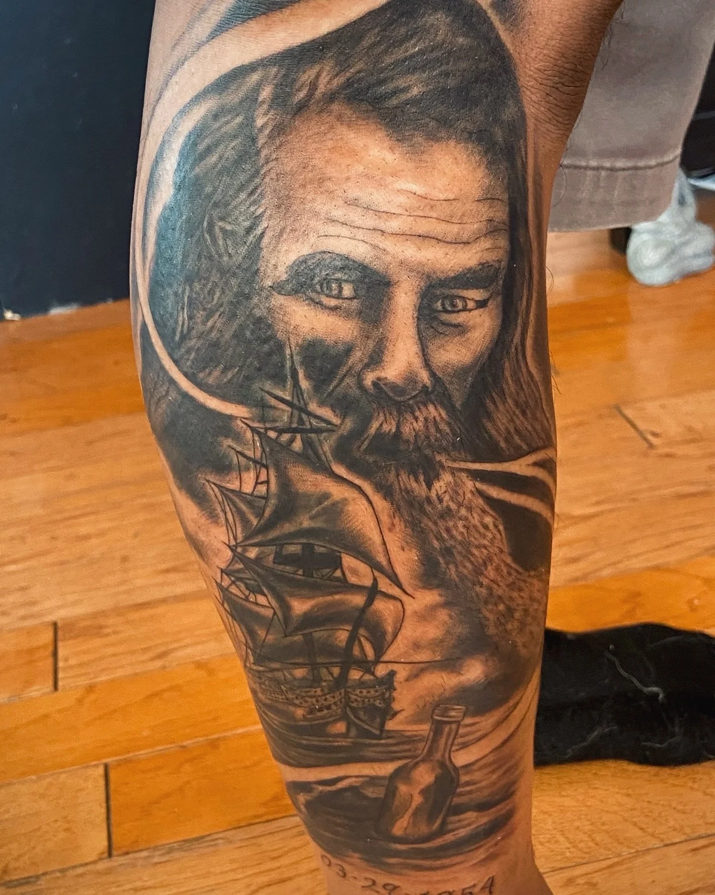 A tattoo of a man's face with long hair and a beard on a person's arm, with a ship sailing on water beneath the face.
