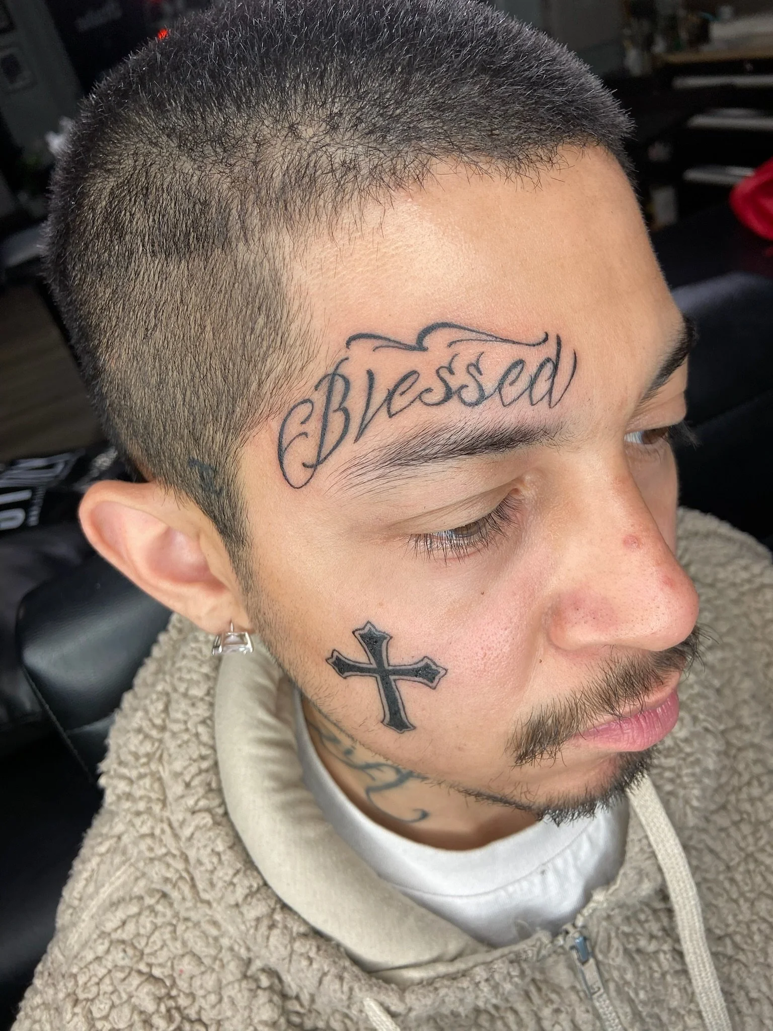 A young man with short dark hair and tan skin, lying down with his eyes closed, showing tattoos on his forehead and cheek, including the word 'Blessed' above his right eyebrow and a cross on his left cheek.