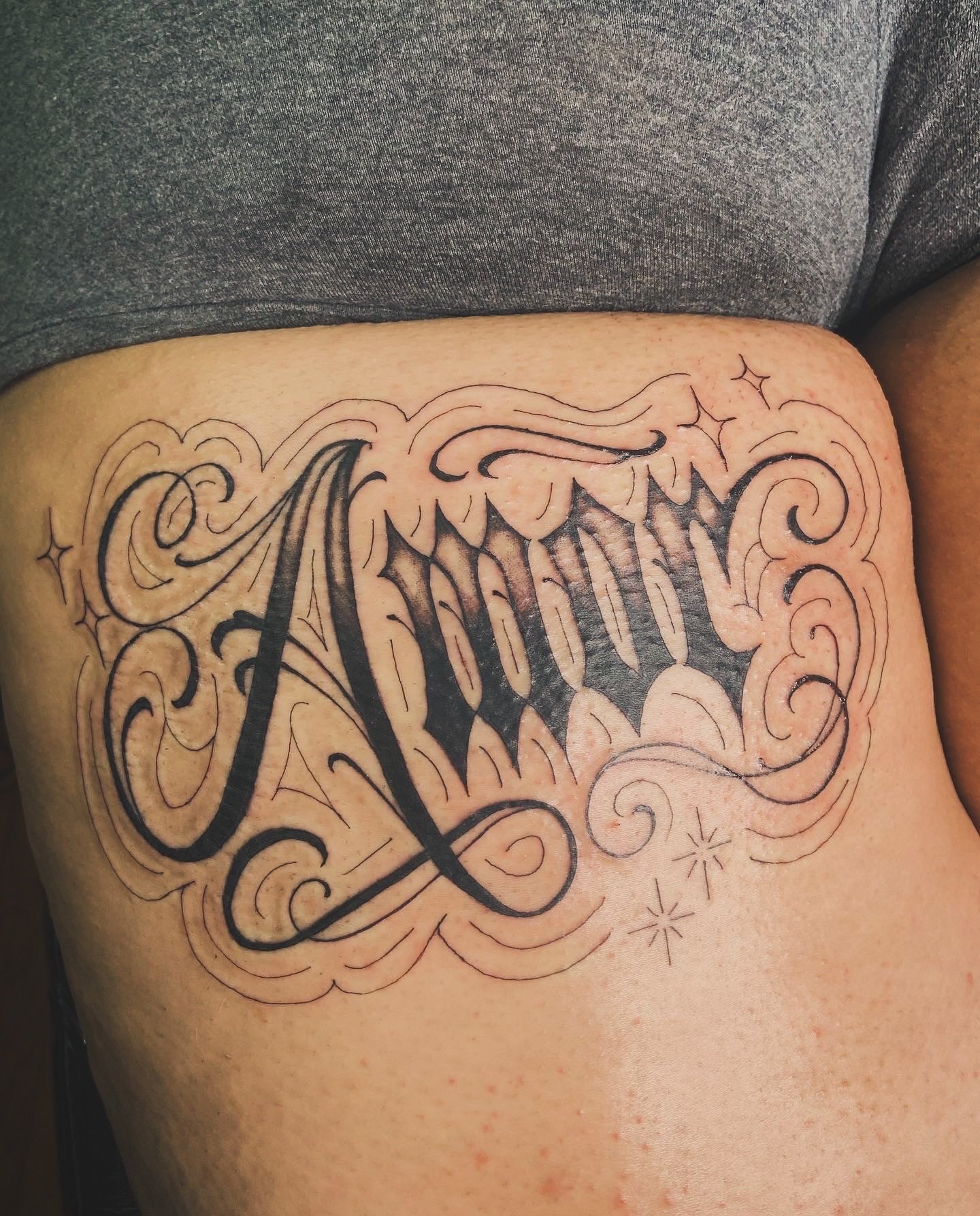 A tattoo on a person's side features the word 'Mom' in elaborate, decorative black script with swirling lines and star-like embellishments.