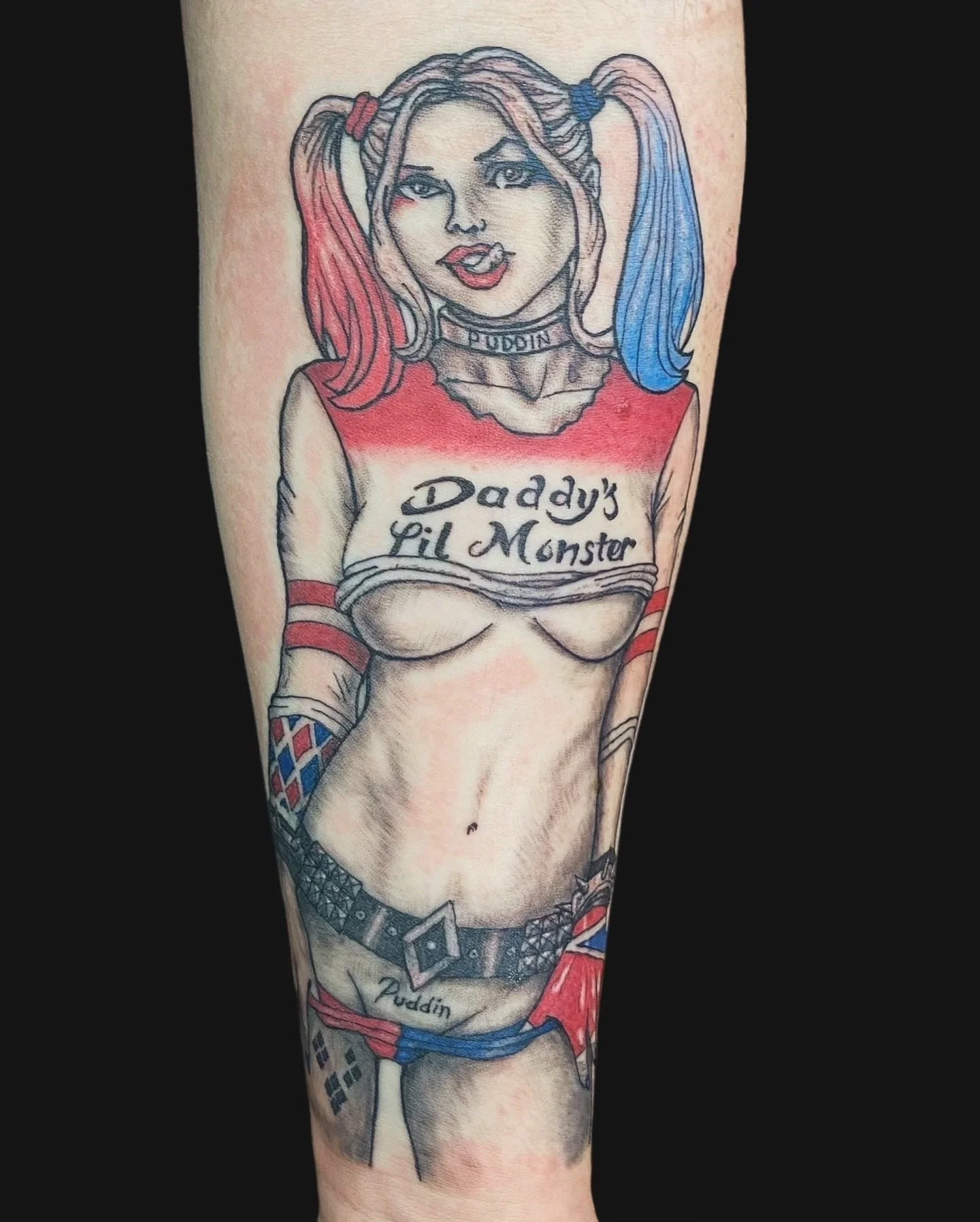 Tattoo of Harley Quinn with blonde hair in pigtails, wearing a red and blue top with the words 'Daddy's Lil Monster,' and various accessories including fishnet stockings and a belt.