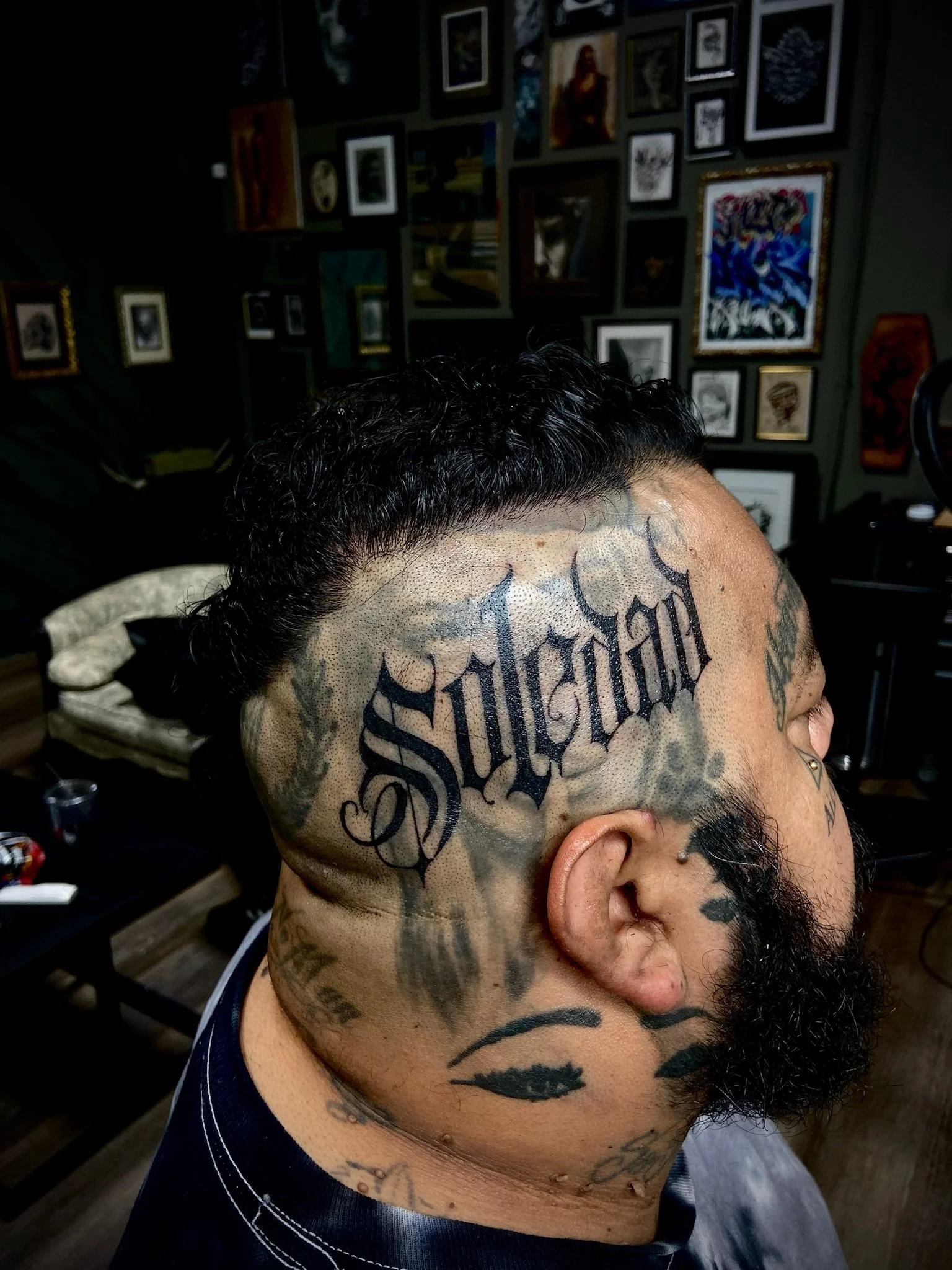 A man with black, curly hair and a beard, showing multiple tattoos on his face, neck, and ear, in a dimly lit room with a gallery of framed pictures on the wall behind him.