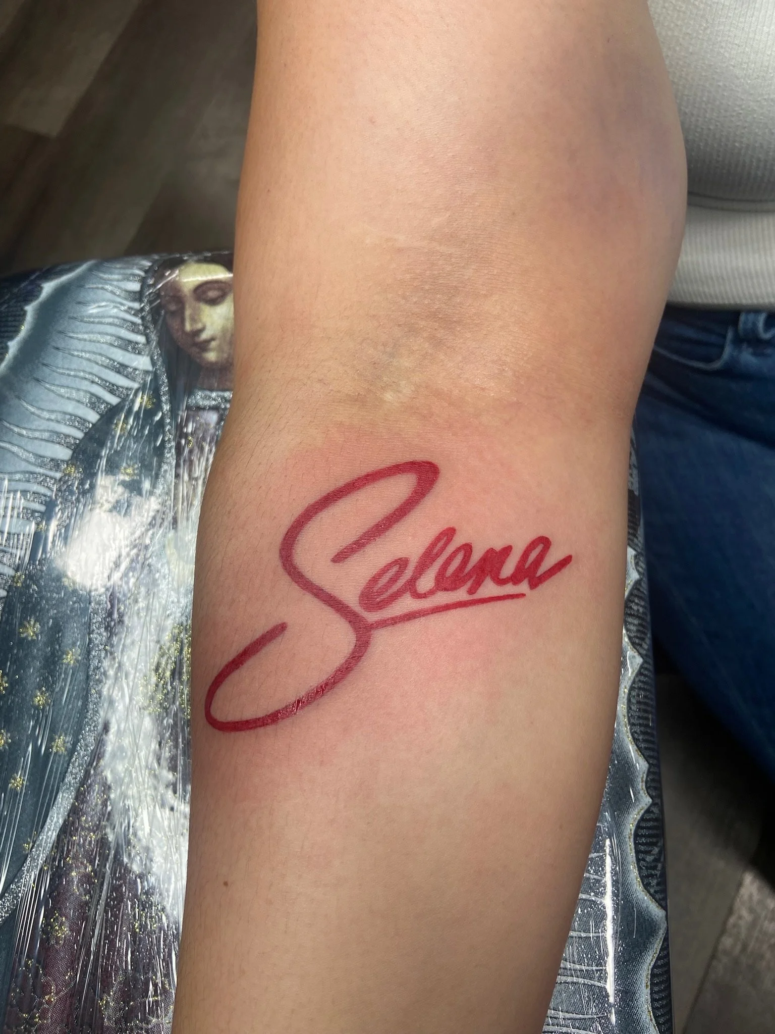 Close-up of a person's arm with a red tattoo reading 'Selena' on the wrist, next to a faint scar.