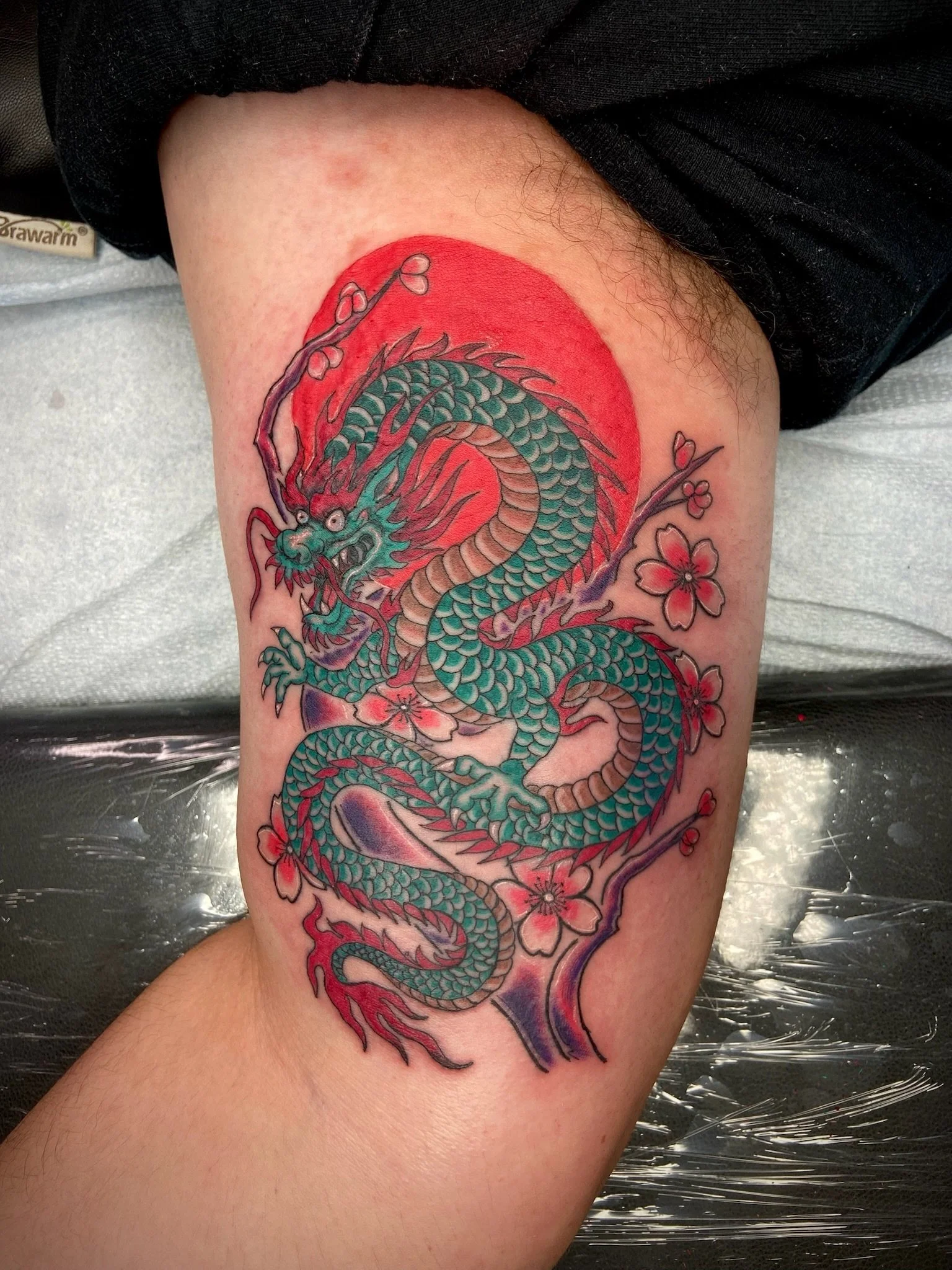 Colorful dragon tattoo on a person's thigh, with a red sun and cherry blossom flowers.