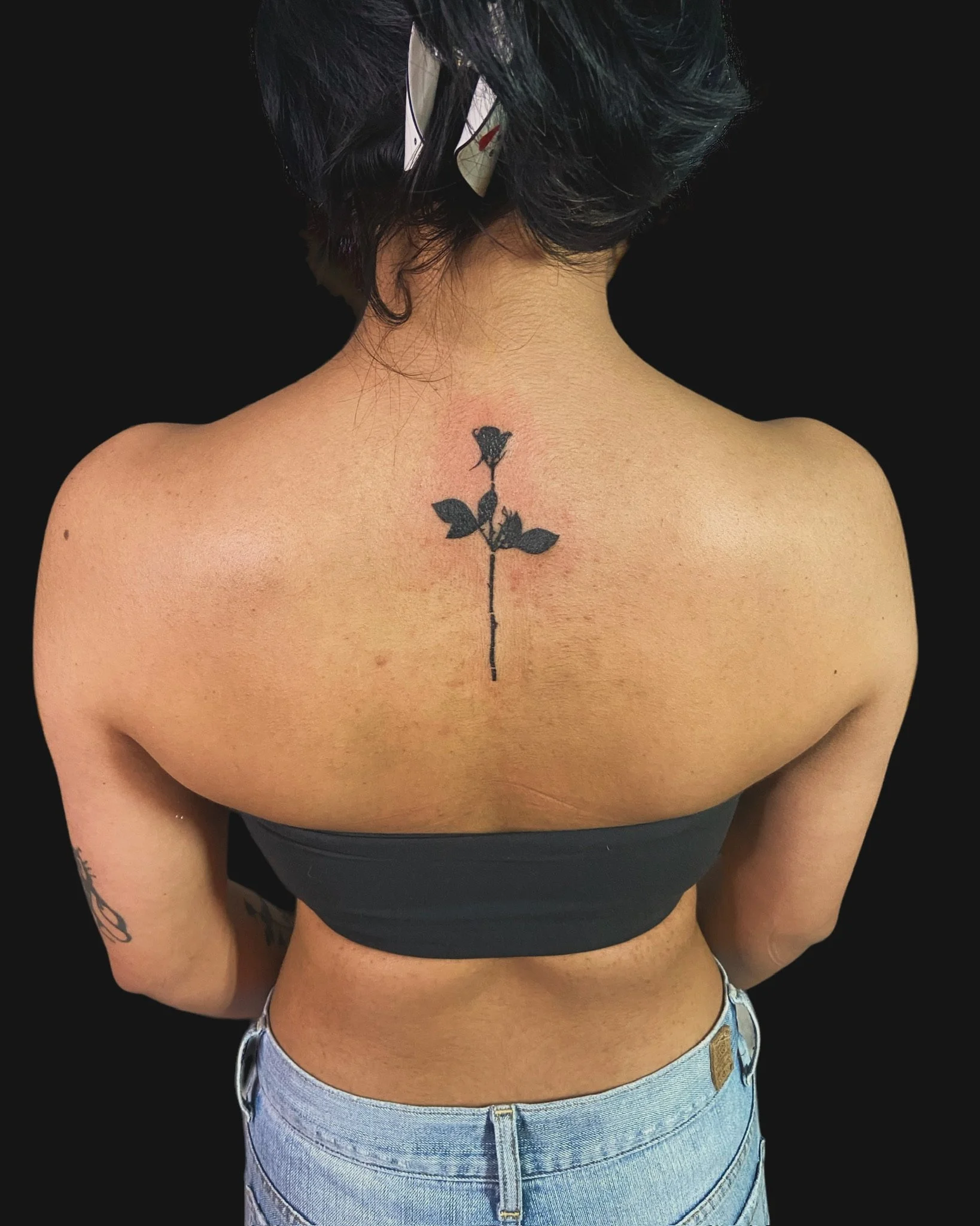 Back view of a woman with curly dark hair, wearing a black top and light blue high-waisted jeans, with a tattoo of a single rose on her upper back between her shoulders.