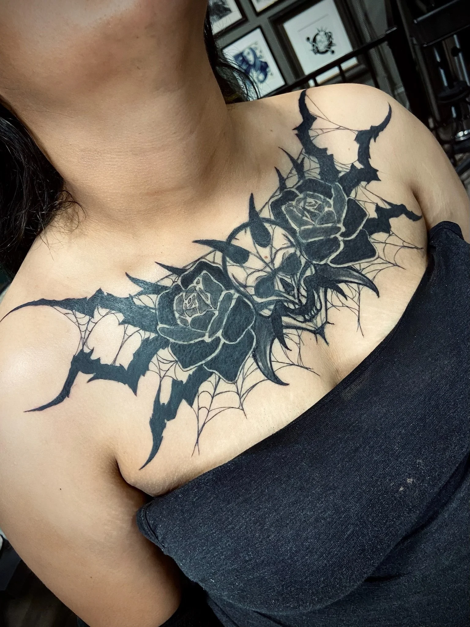 Close-up of a person's shoulder and collarbone with a large black tattoo of a rose surrounded by jagged, thorny vines and intricate web-like details.