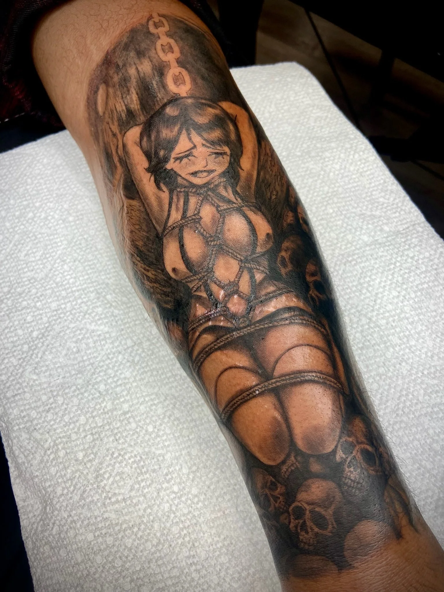 A detailed tattoo on a person’s arm featuring a cartoonish girl with dark hair and a distressed facial expression, with background elements including a chain pattern and abstract designs in shades of black and gray.
