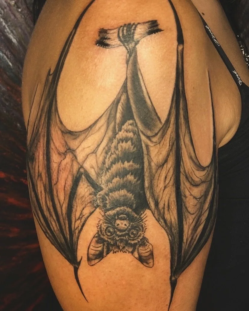 Tattoo of a bat with detailed wings and a bee's face on its body on a person's upper arm.