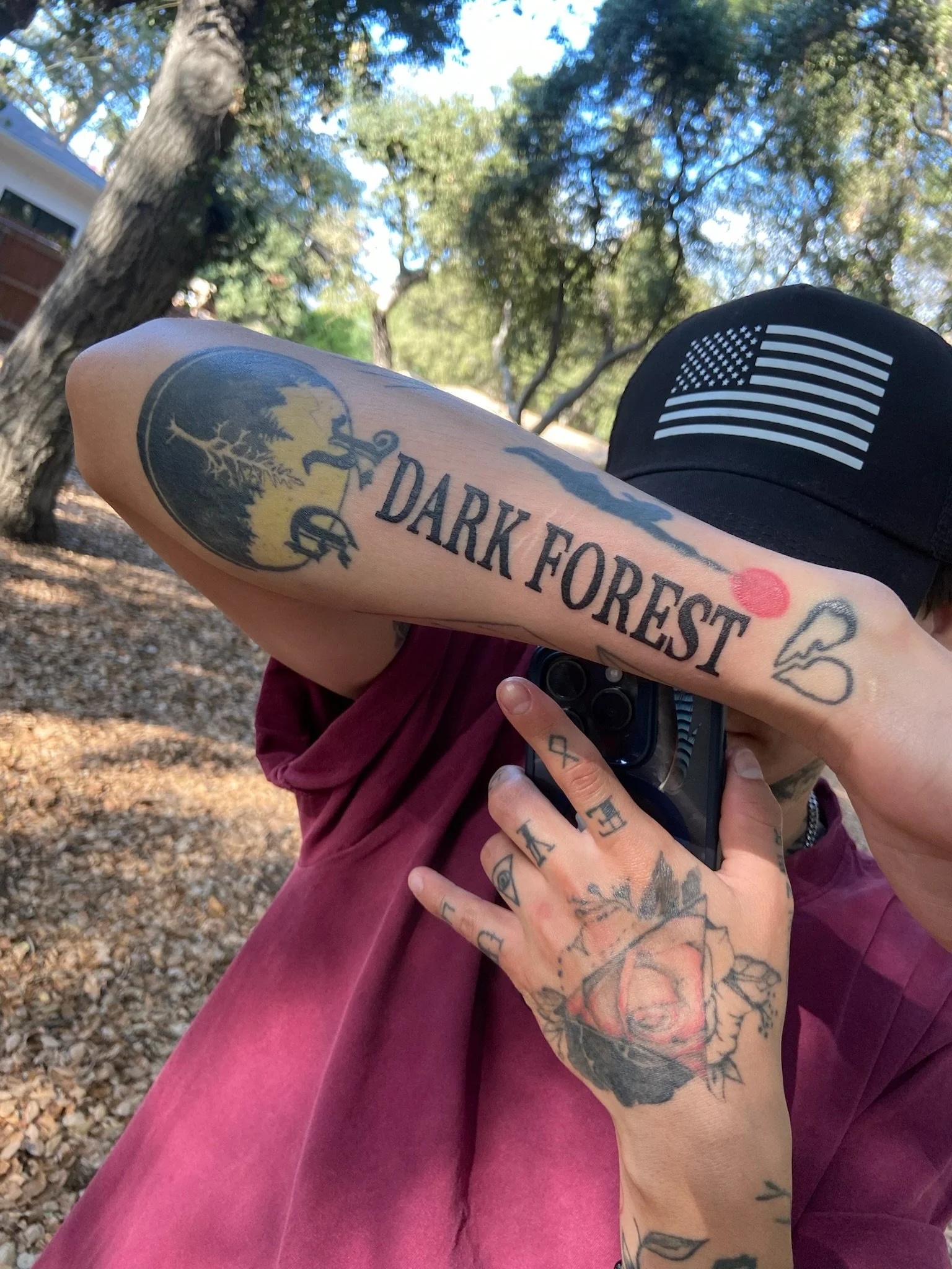 Close-up of a person's arm with tattoos, holding a smartphone, in front of a black cap with an American flag patch, outdoors among trees.