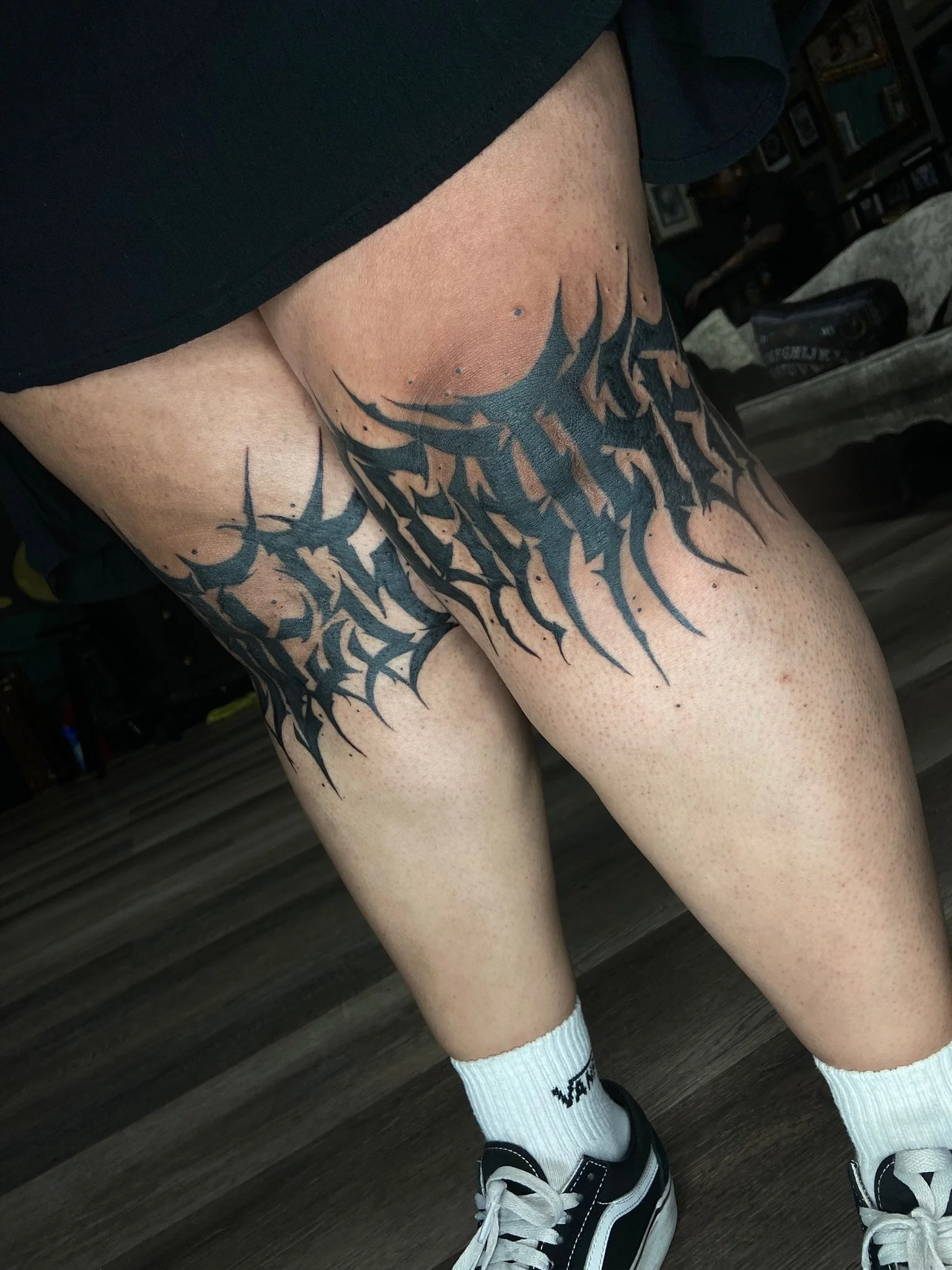 A person's legs wearing black and white Vans sneakers, white calf-high socks, and black shorts. The person has a large black and orange abstract tattoo across their thighs, with a background of a wooden floor and some furniture.