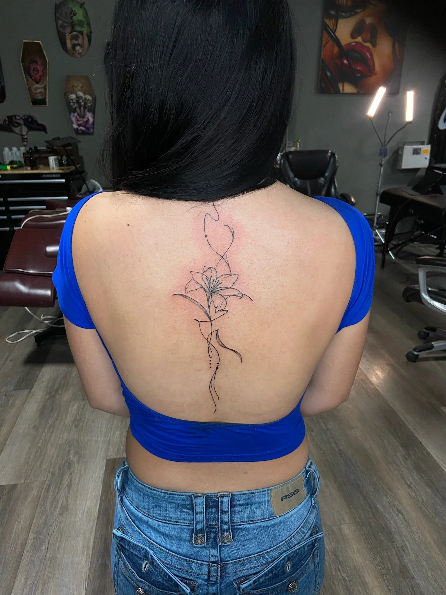 Back view of a woman with a large tattoo of a flower and abstract design on her upper back, wearing a blue top and jeans, in an art studio or tattoo parlor.