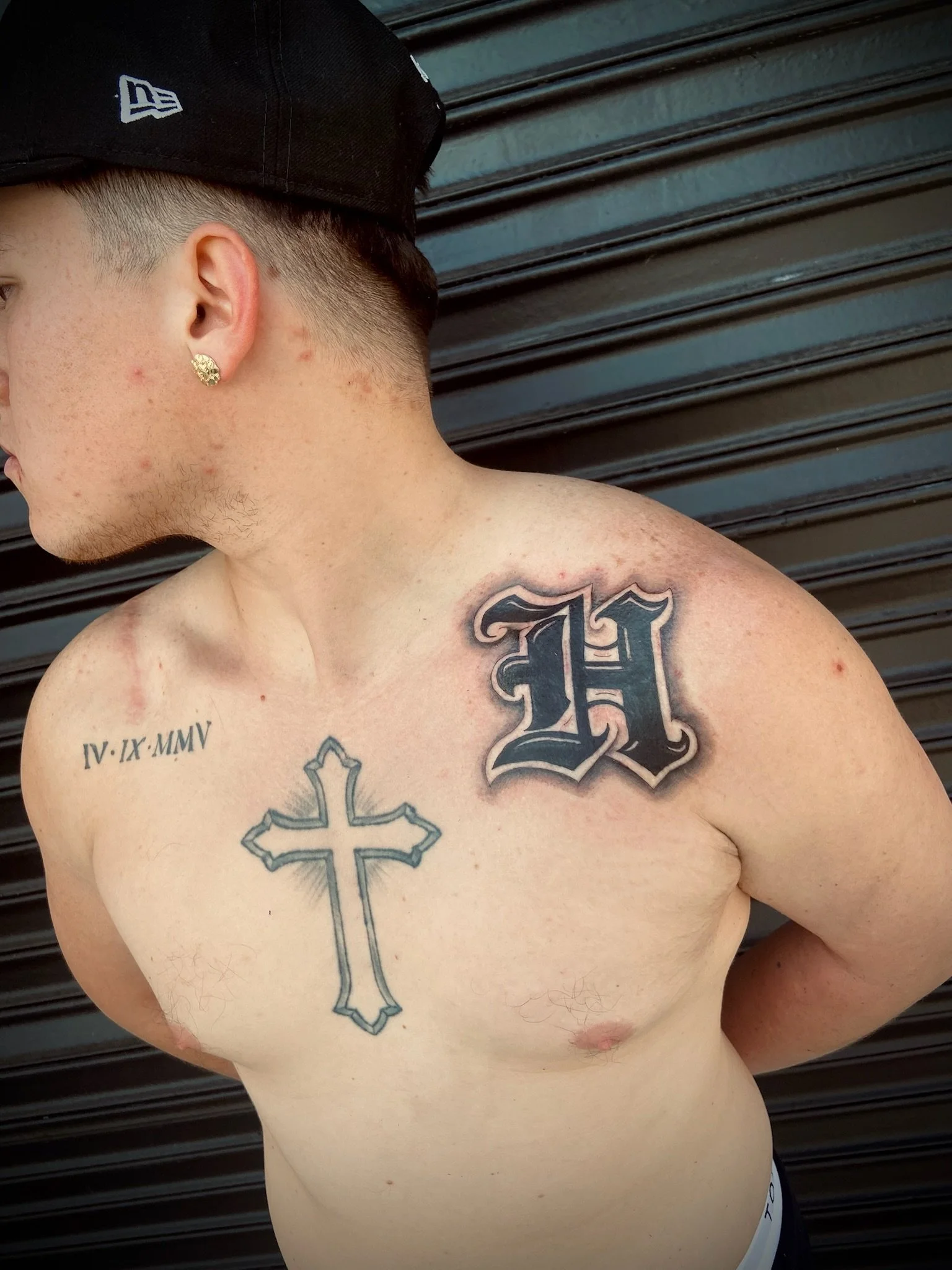 A shirtless young man wearing a black baseball cap and gold earrings, with tattoos on his shoulder and chest, standing in front of a dark metal wall.