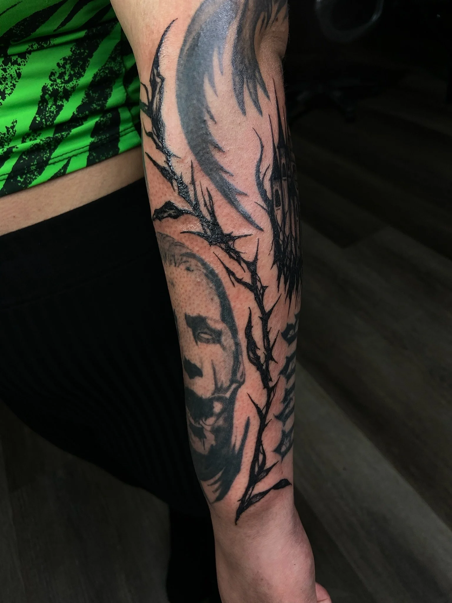 Close-up of a person’s arm with a large black and gray tattoo of a tree with branches extending down the arm, next to a green shirt with black patterns.