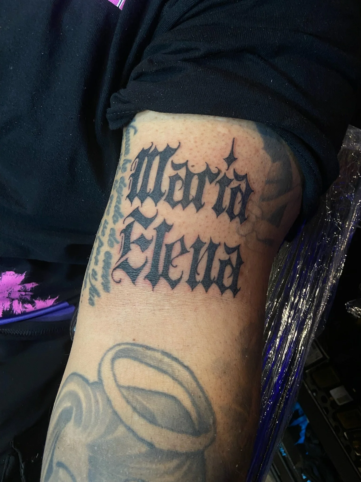 Close-up of a person's arm with black tattoo text and patterns, wearing dark clothing, next to someone sitting on a chair with part of a colorful garment visible.