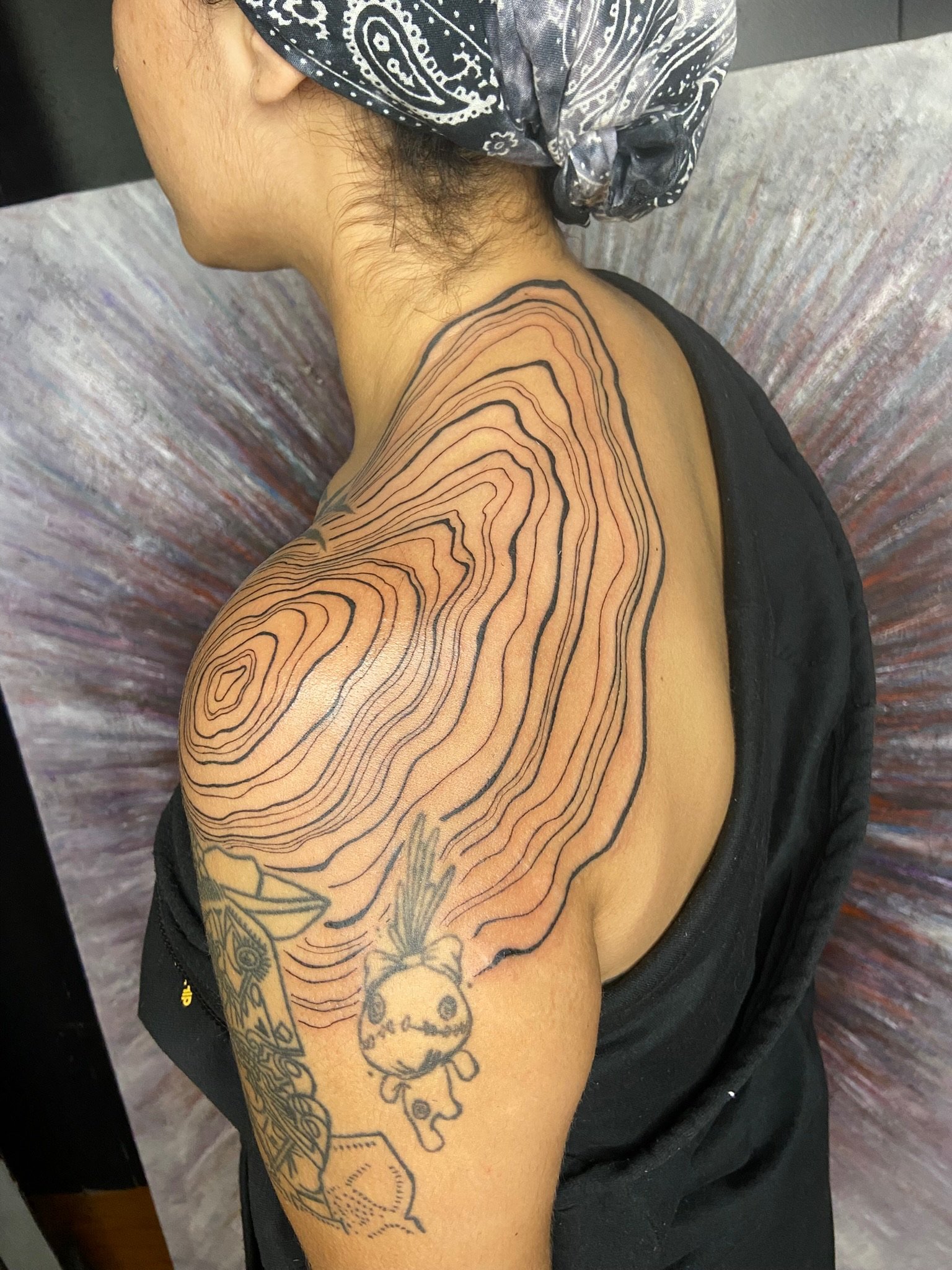 A person's upper arm with a tattoo of a butterfly, a skull, and contour lines, seated on a textured, multicolored surface, wearing a dark bandana with a white paisley pattern.