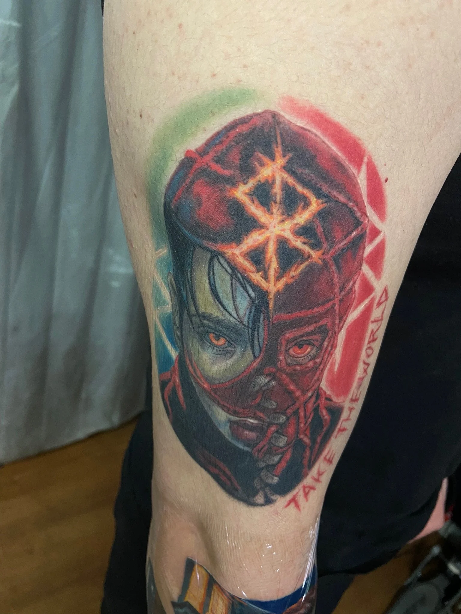 Tattoo of a human face with dark hair and red eyes, featuring a glowing geometric shape on the forehead and red surrounding marks, with red lettering on the side.