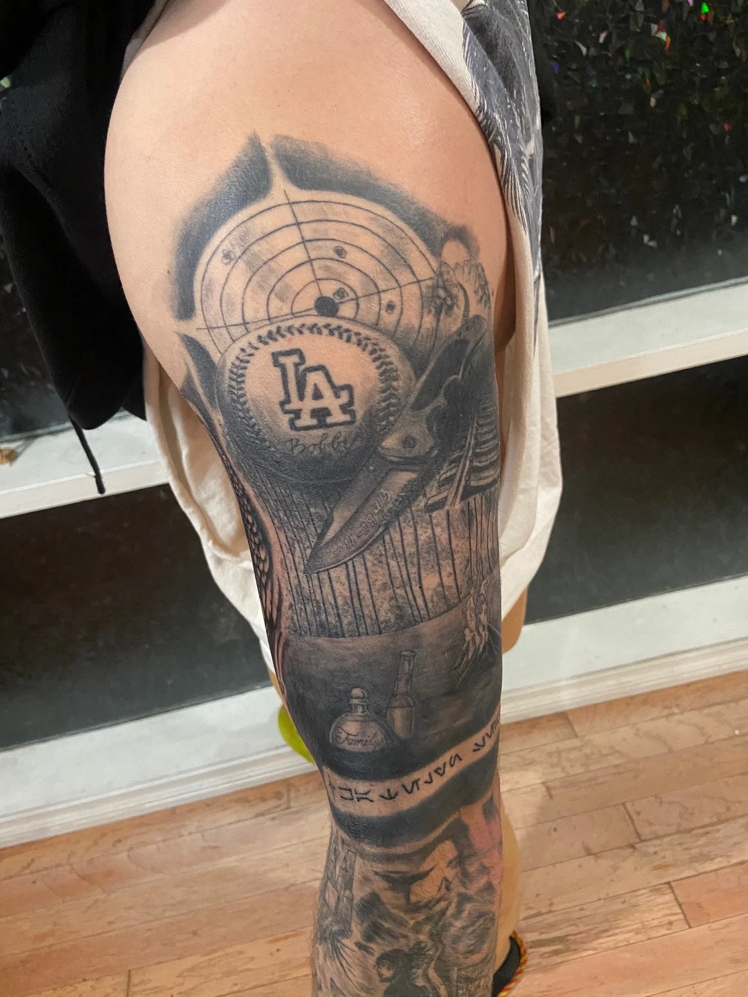 A person's arm with a tattoo featuring a dartboard, the Los Angeles Dodgers logo, a sword, and various other designs.