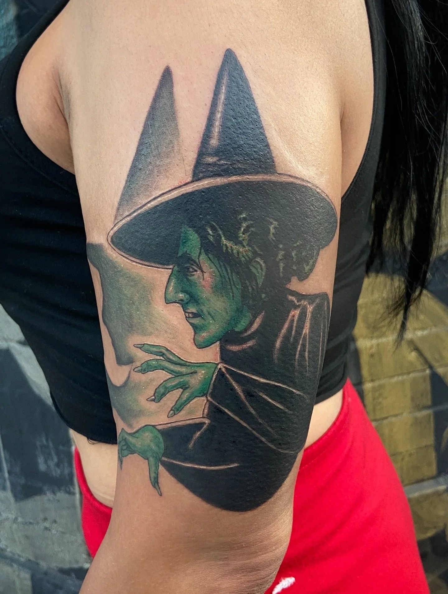 Tattoo of a woman with green skin, wearing a black witch's hat, on a person's upper arm.
