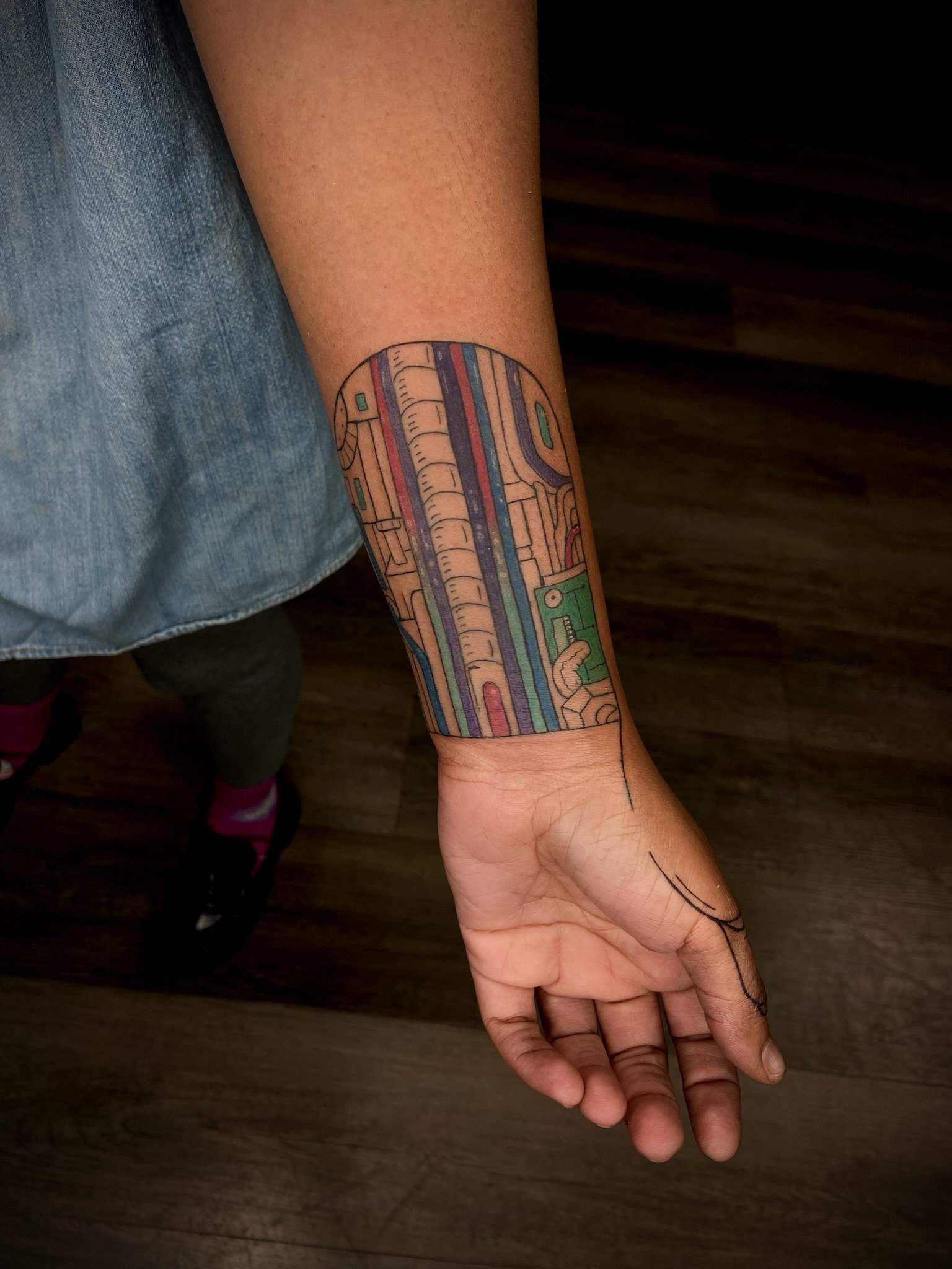 A person's left arm extended downward, showing a colorful tattoo sleeve with intricate geometric and abstract patterns.