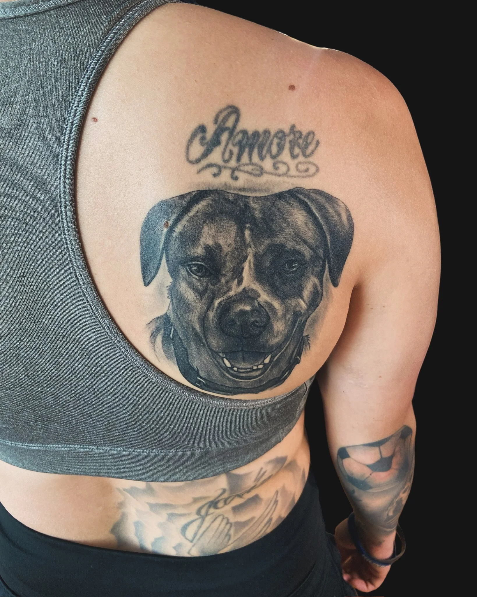 Close-up of a tattoo on a person's upper chest featuring the word 'Amore' written in cursive, with a puppy dog tattoo below it, and part of their arm and side in view.