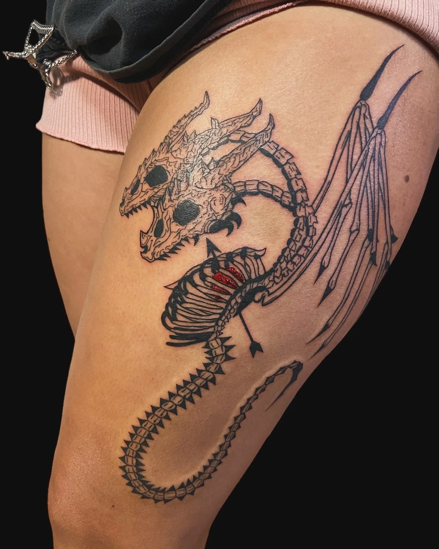 Black tattoo of a dragon skeleton on person's upper thigh.