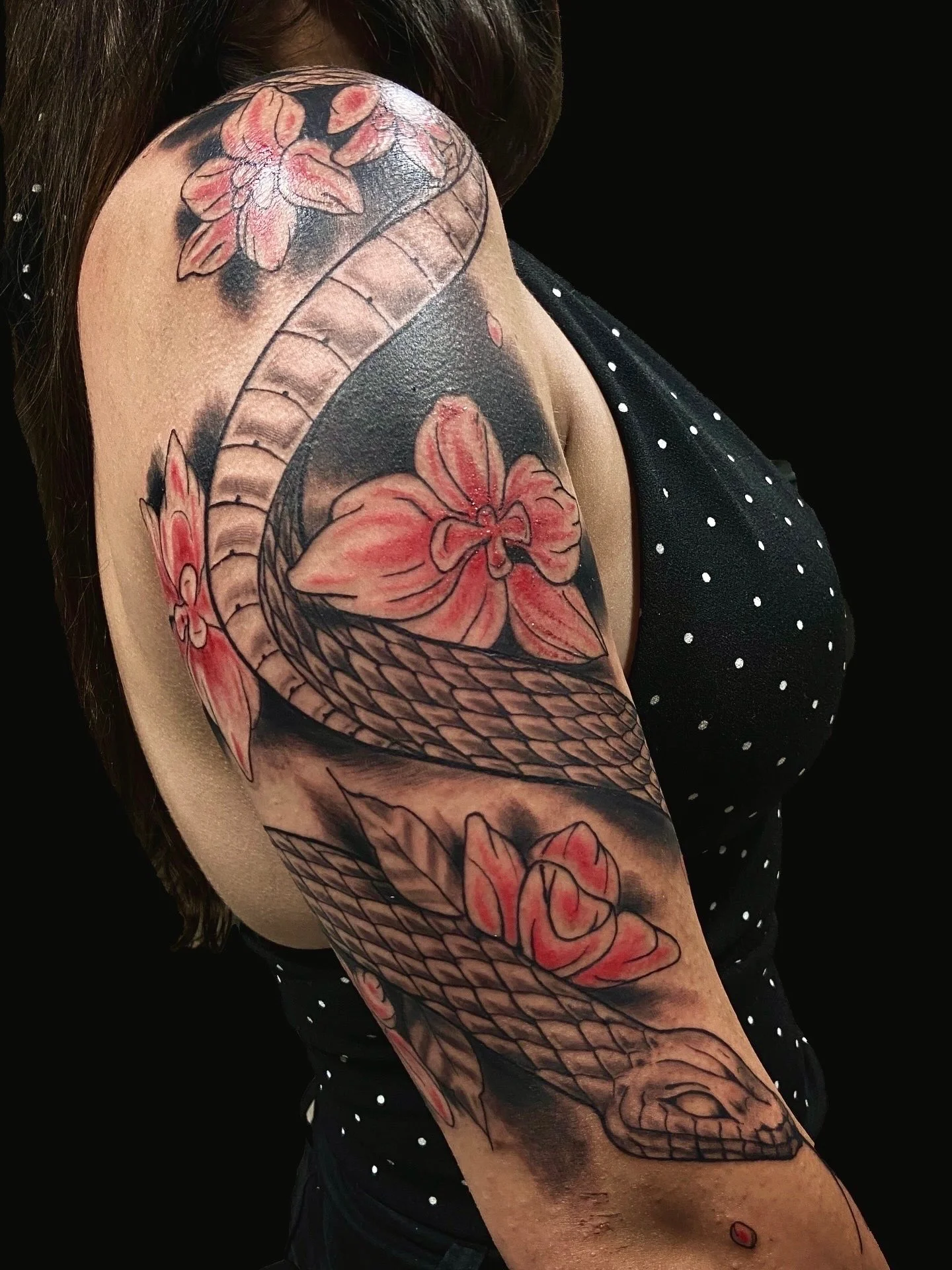 A person's upper arm with a tattoo of a snake intertwined with pink flowers and leaves, against a black background.