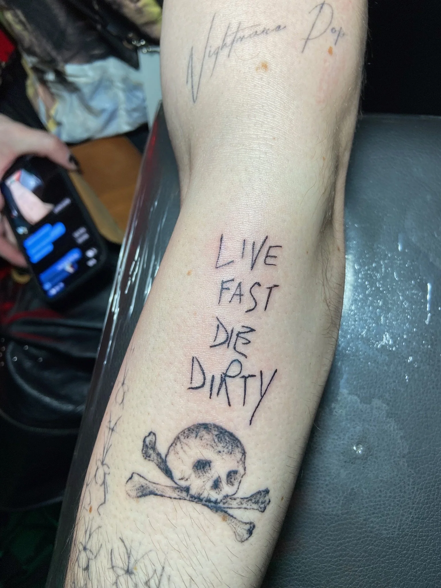 Tattoo of a skull with crossbones on a person's forearm, with handwritten words 'Live Fast Die' and other words partially visible.