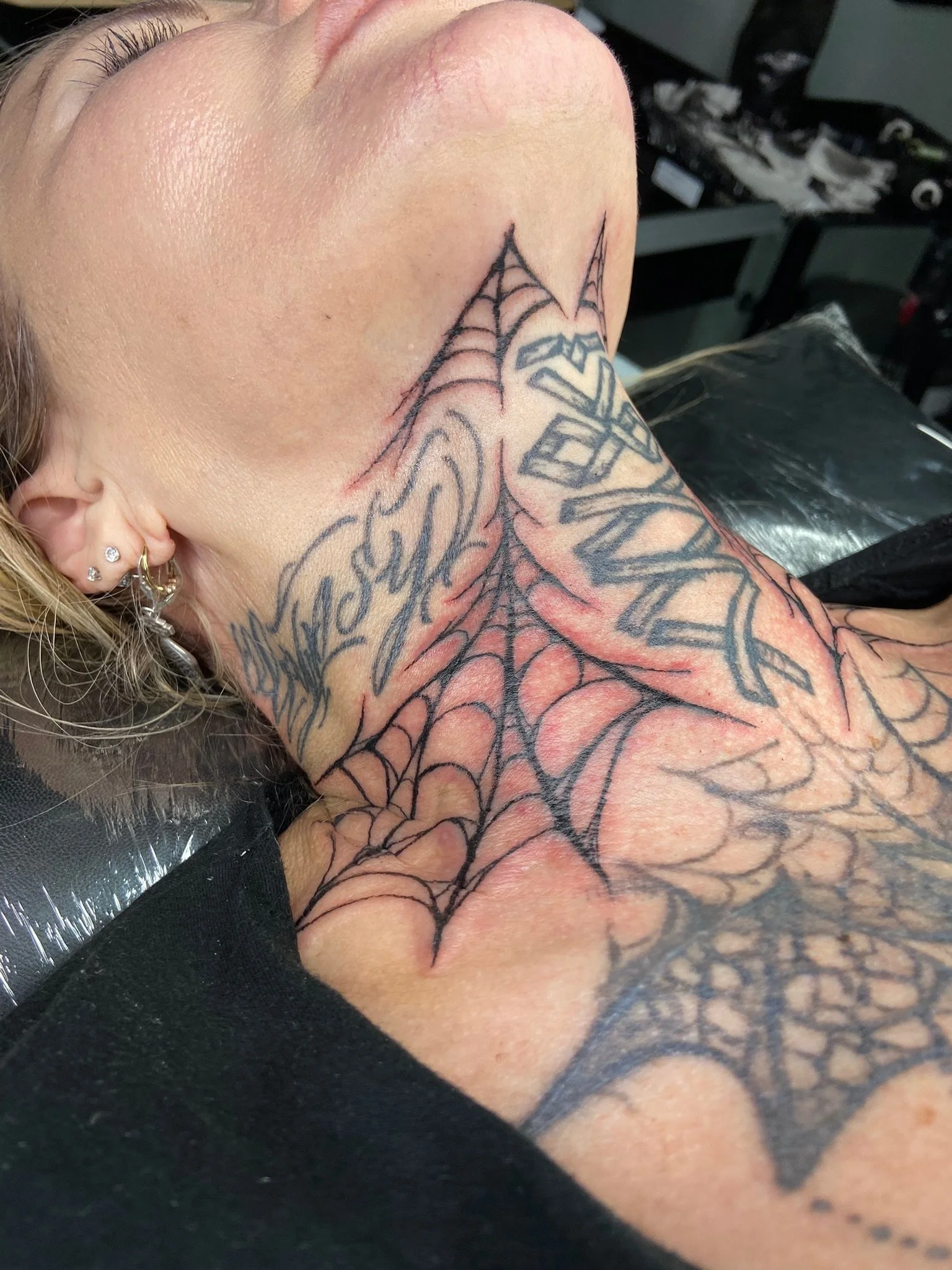 Close-up of a person's shoulder and neck with a spider web tattoo and text tattoo, wearing earrings.