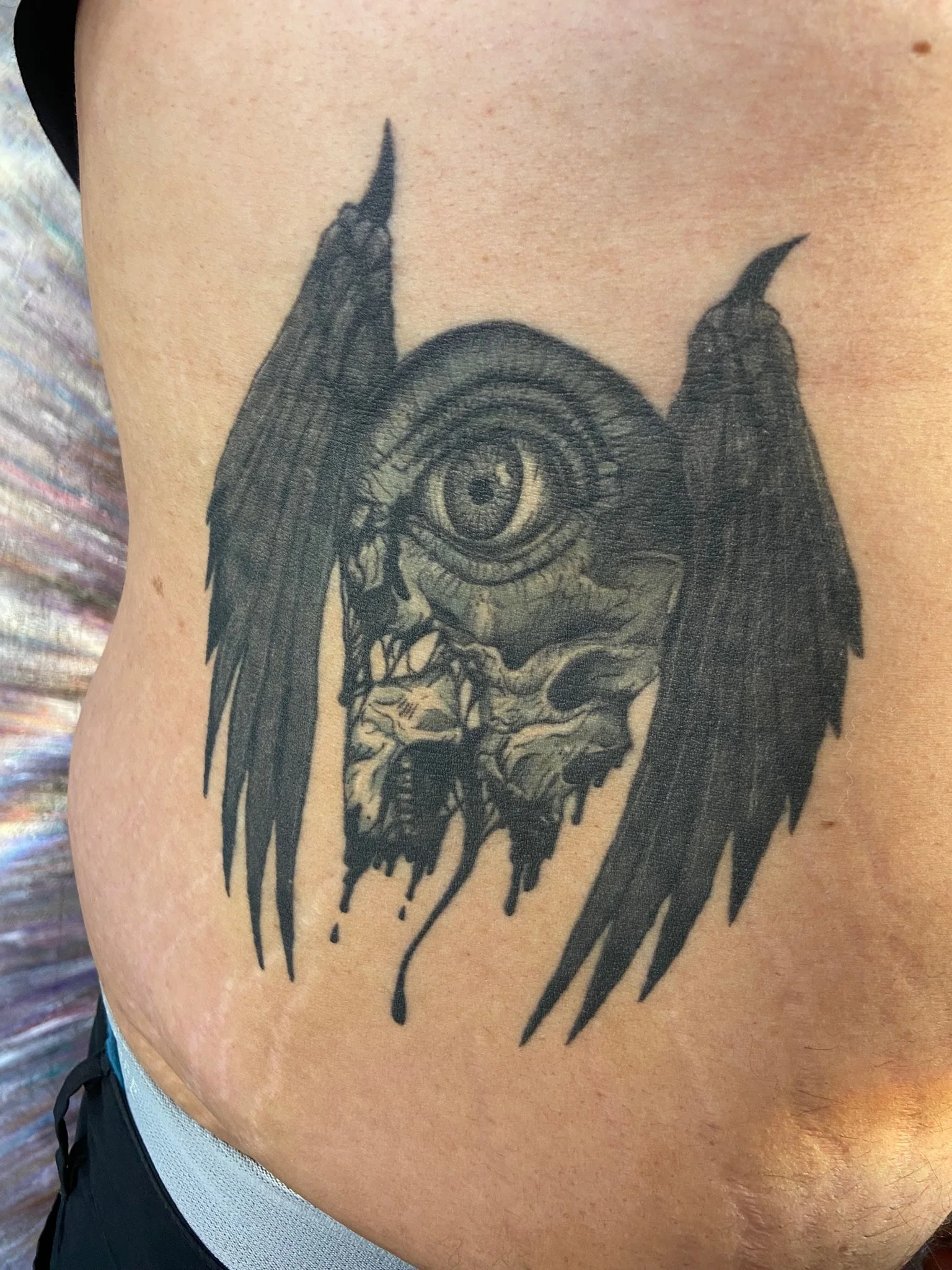 A black tattoo on a person's side of a dripping skull with an eye and wings.