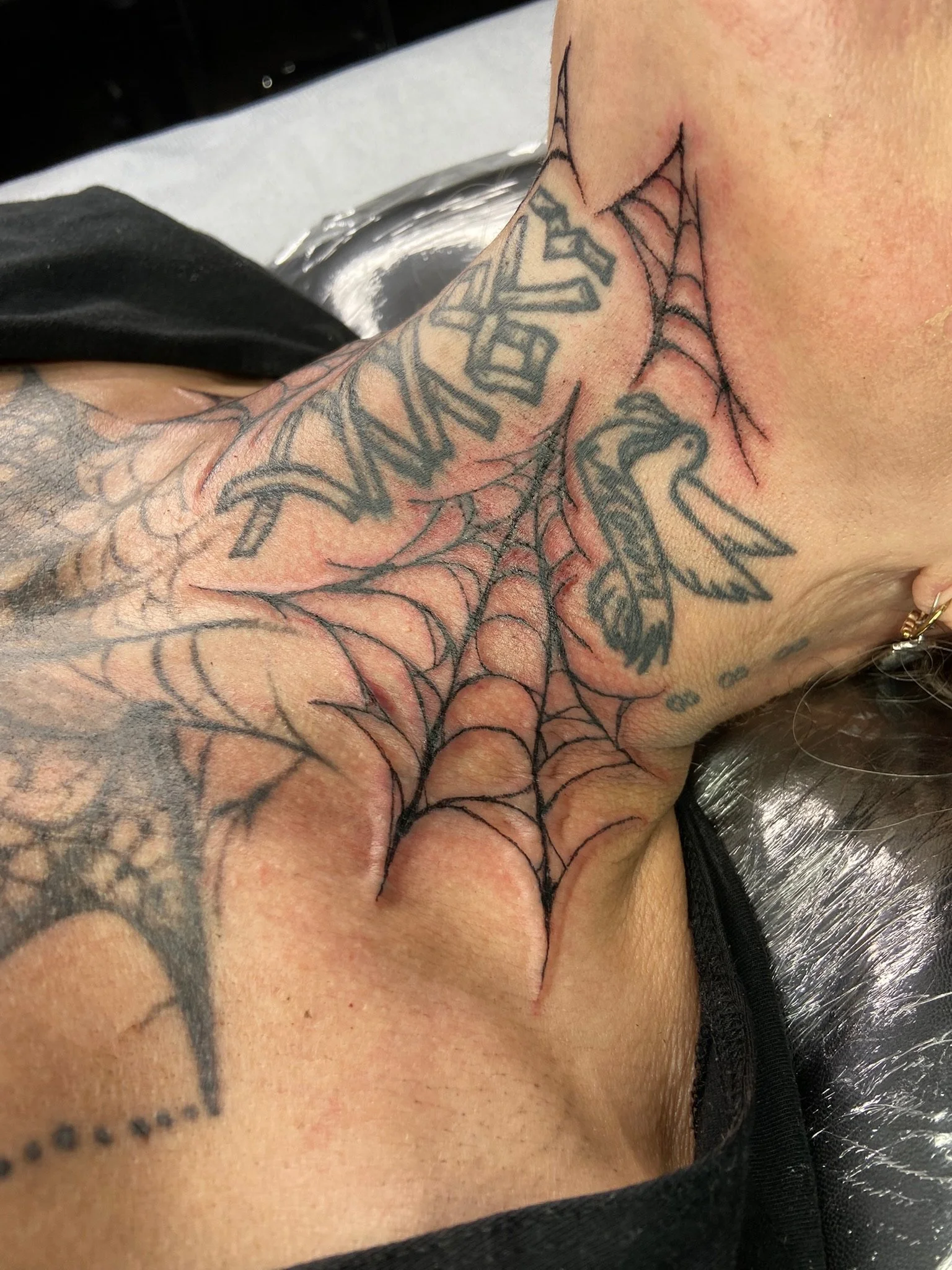 Close-up of a tattoo on a person's neck and shoulder, featuring a spider web and lettering.