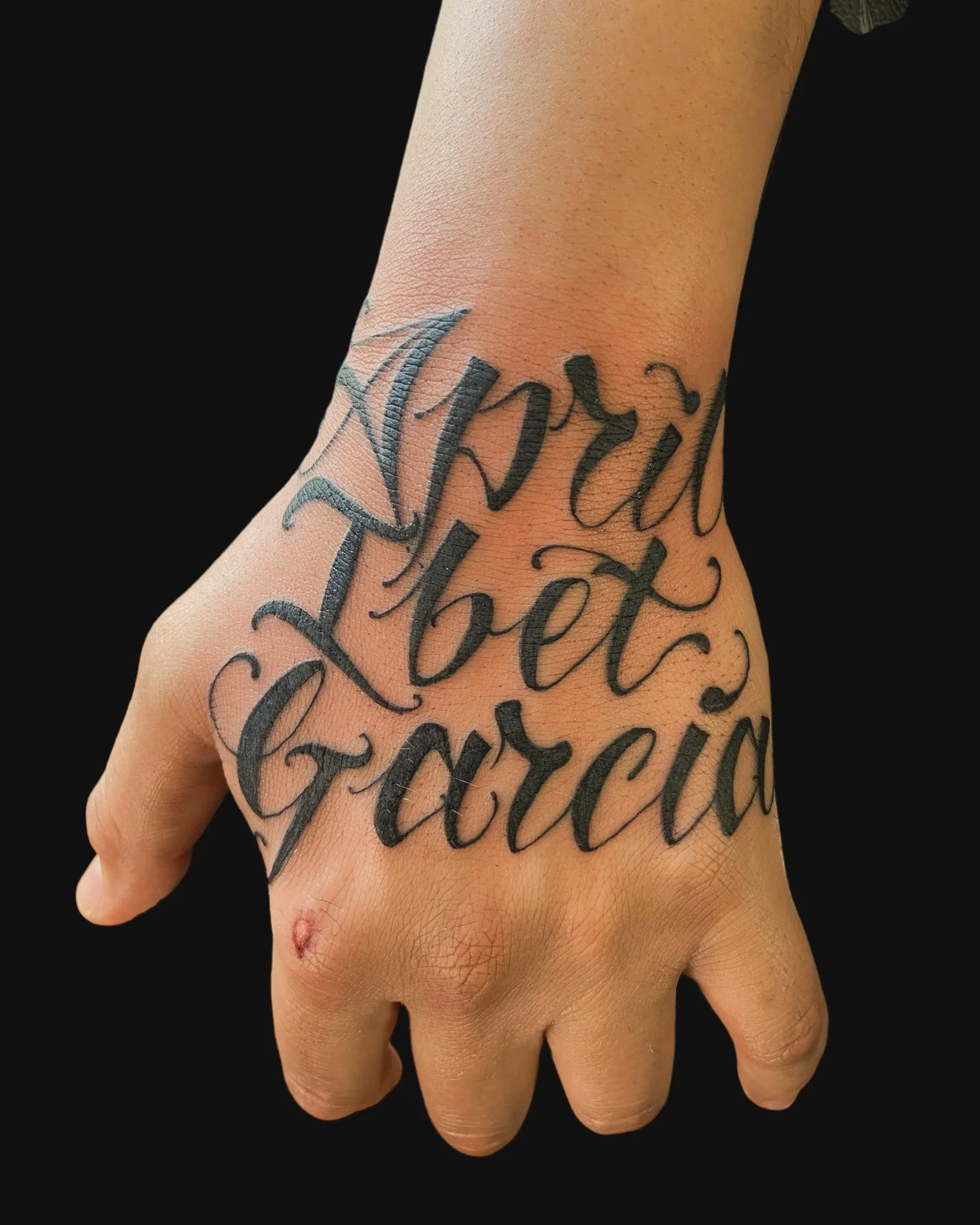 Close-up of a person's hand with a tattoo of cursive text on the wrist and lower forearm, and a small healing wound on the thumb.