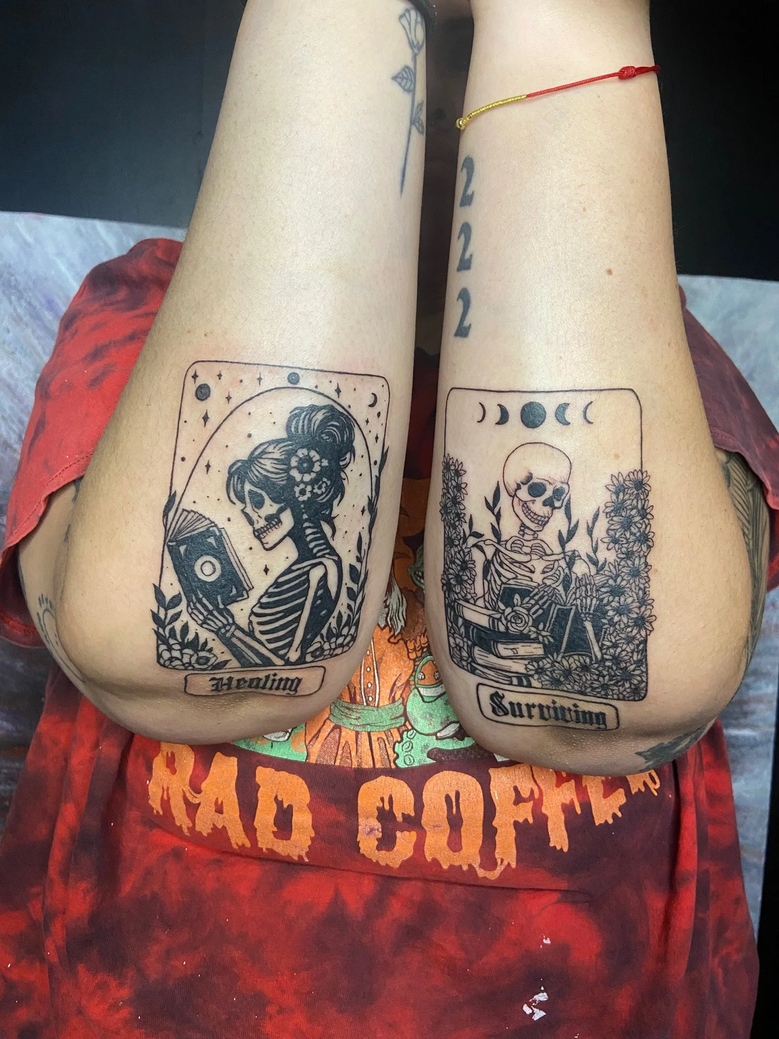 Person with backwork tattoos on their arms. The left arm has a tarot card tattoo labeled 'Healing,' depicting a woman with flowers in her hair. The right arm has a tarot card tattoo labeled 'Surviving,' showing a skeleton with a skull and bones.