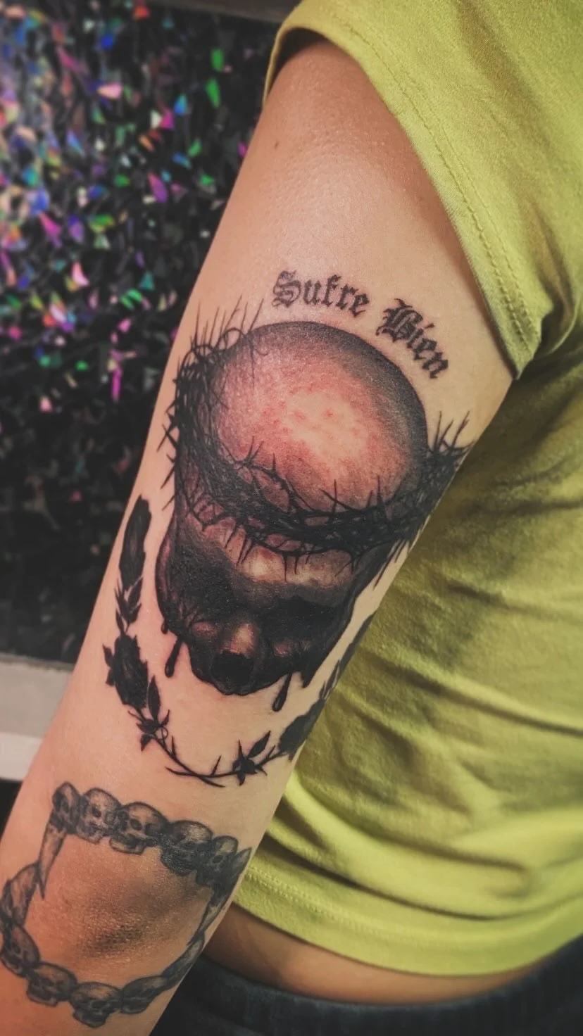 Tattoo of a skull with a crown of thorns, with the words 'Suffer Well' above it, on a person's arm.