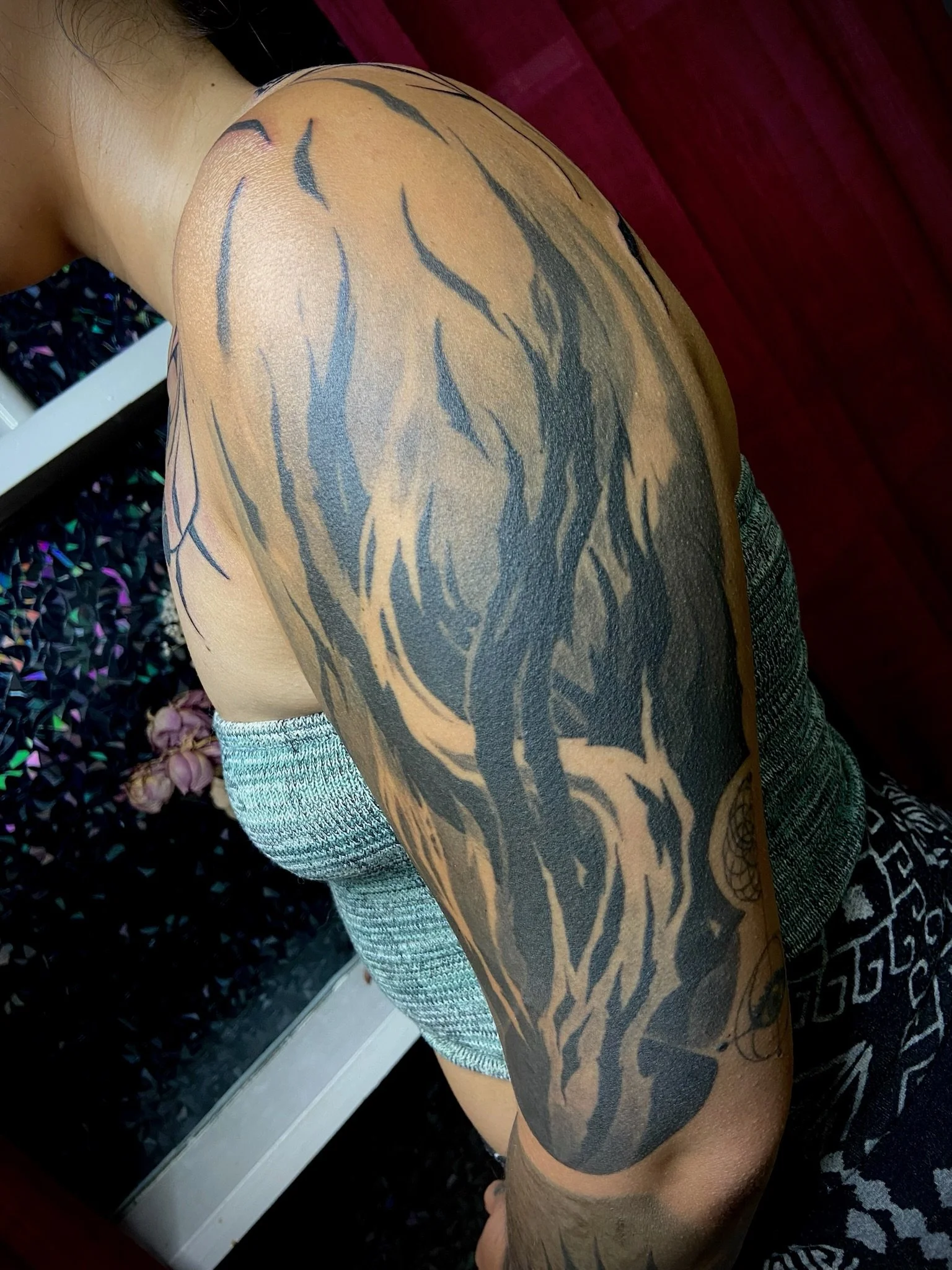 Close-up of a person's shoulder and upper arm with a large black and gray tattoo of a horse's head. The person is wearing a gray short-sleeved shirt with a textured pattern. The background includes a dark red piece of furniture and a black surface wi