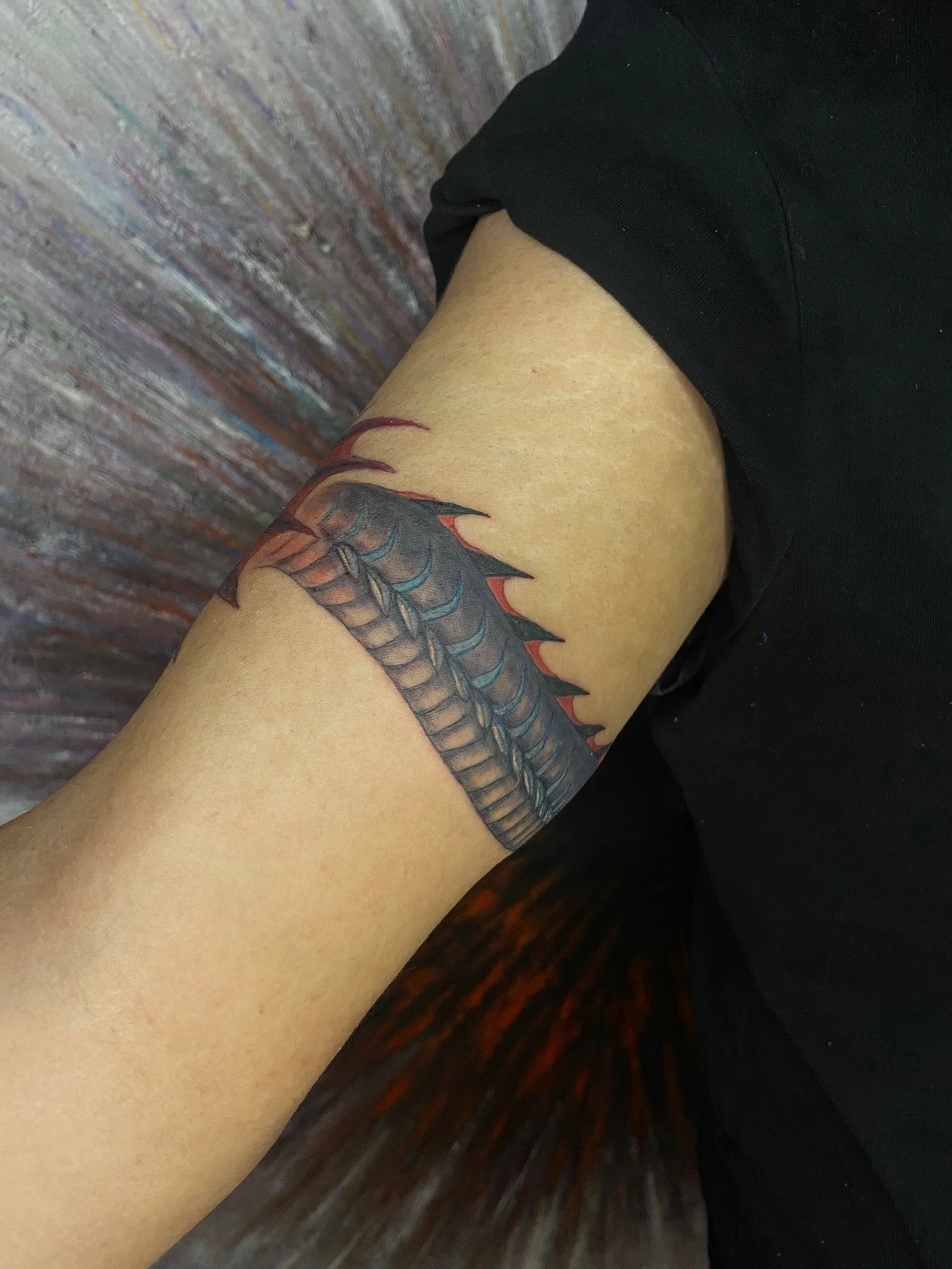 Close-up of a person's upper arm with a colorful dragon tattoo, sitting next to a textured brick wall and wearing a black shirt.
