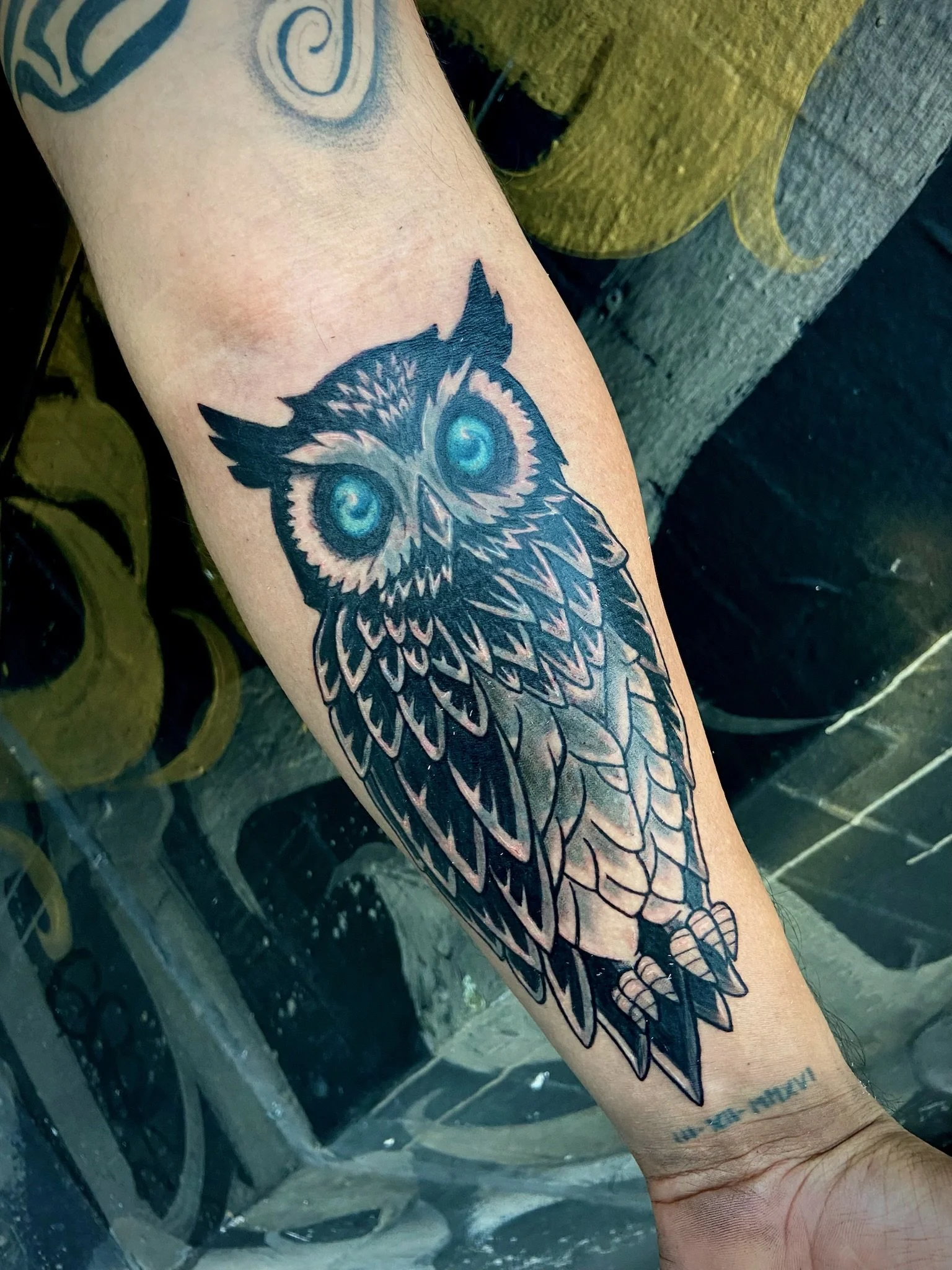 Tattoo of an owl on a person's forearm with intricate line work and blue eyes, partially visible with other tattoos in the background.