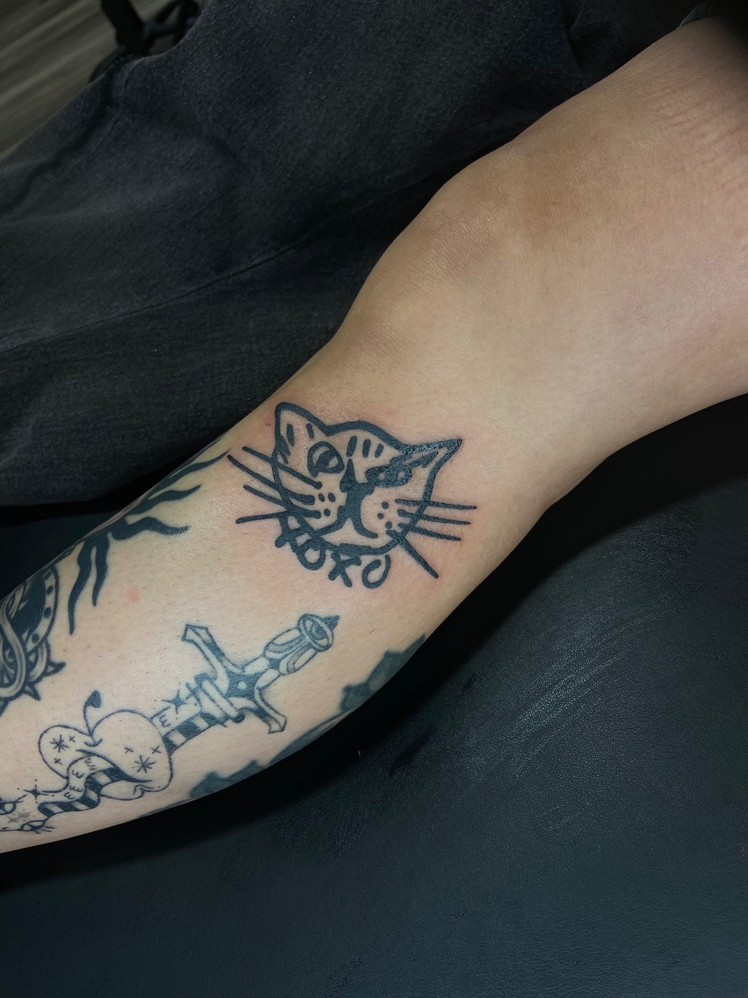 Close-up of a person's forearm with a tattoo of a cat's face and some other designs, resting on dark furniture.