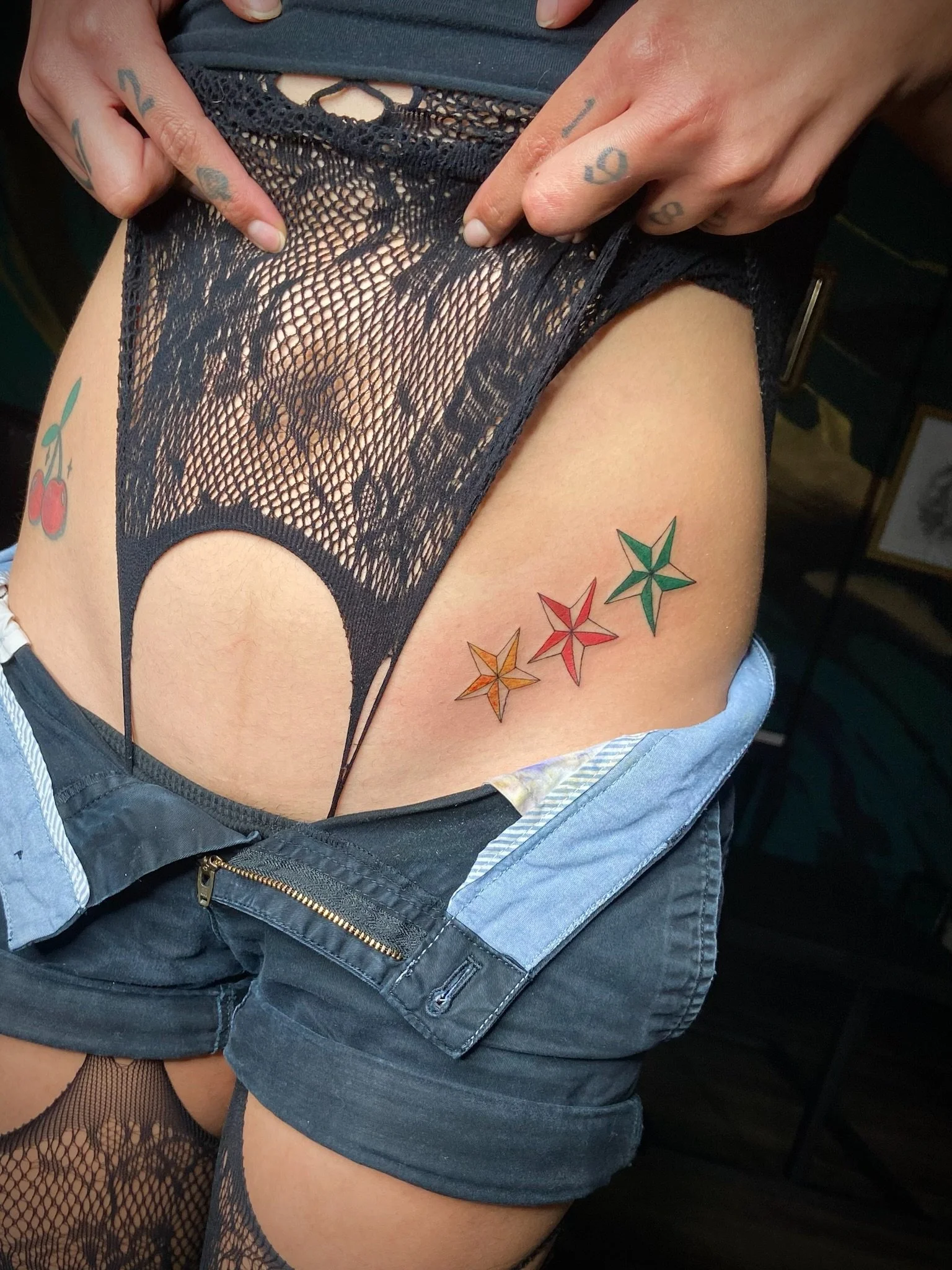 A person wearing black fishnet stockings and dark denim shorts. They are lifting their shirt to reveal a tattooed thigh with a colorful geometric star design.
