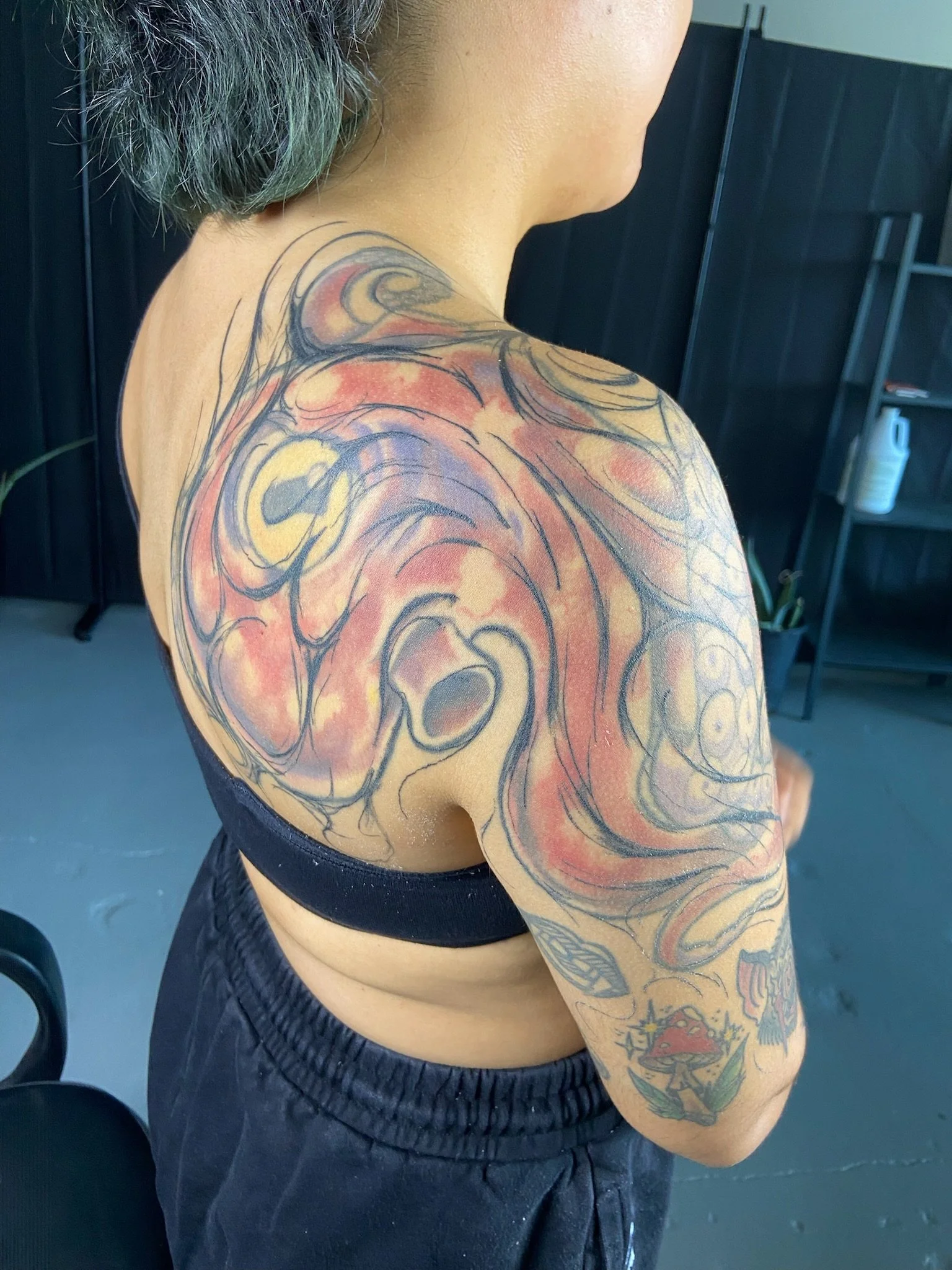 Side view of a person with tattoos on shoulder and arm, wearing a black sleeveless top and black pants, standing in front of a black wall with shelving and a bottle on it. This is the before photo for a coverup tattoo. 