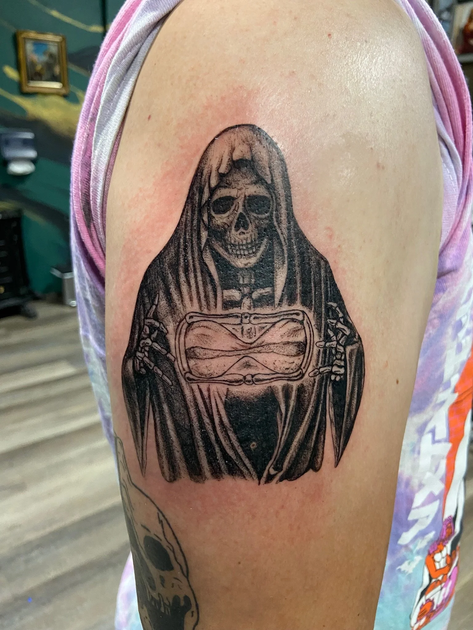 Tattoo of a skeleton with a skull, partially covered by a hood, on a person's upper arm.