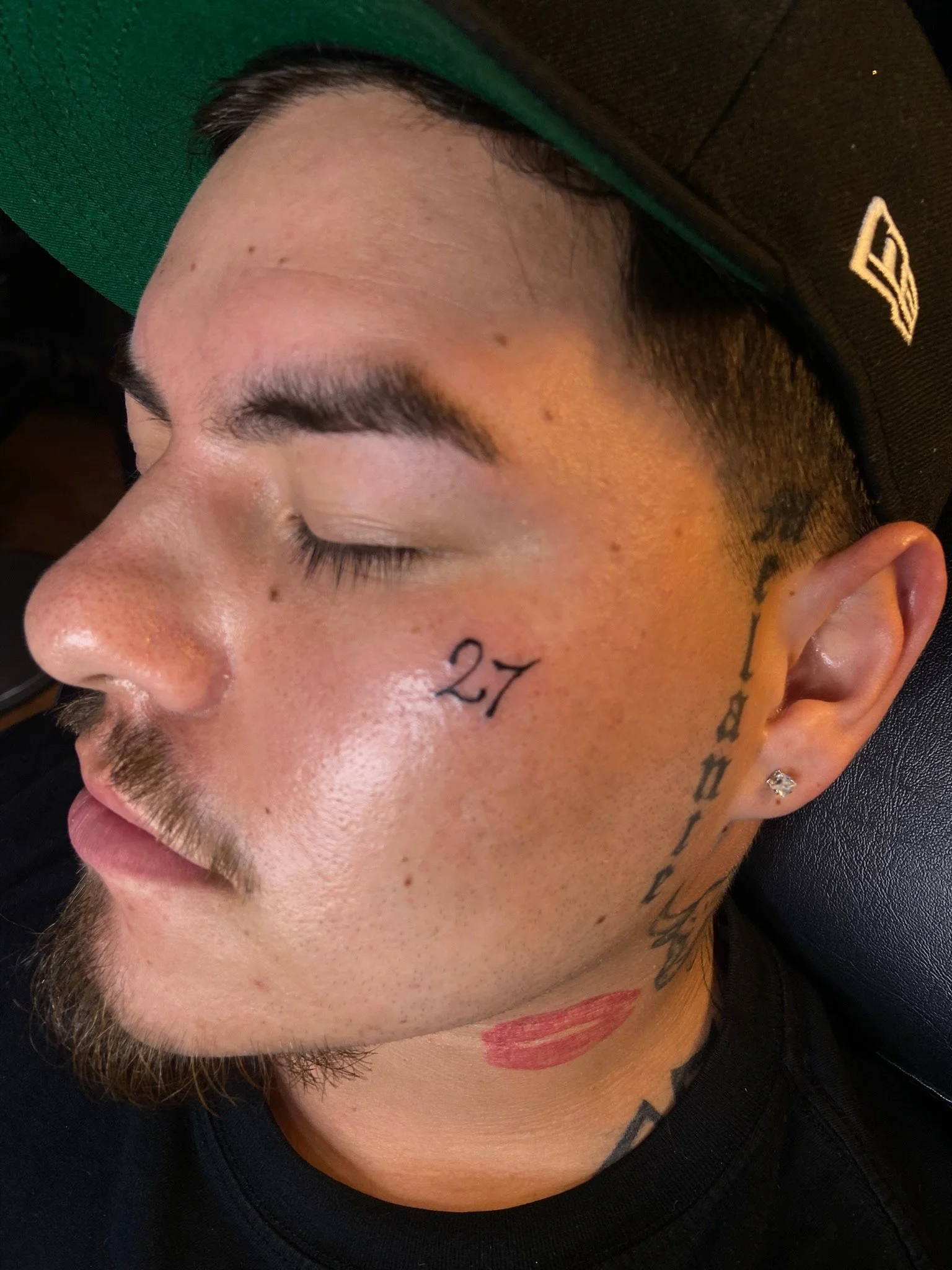 Close-up of a person's face with closed eyes, tattoos on the face and neck, and a colorful lipstick mark on the neck, wearing a black and green cap.