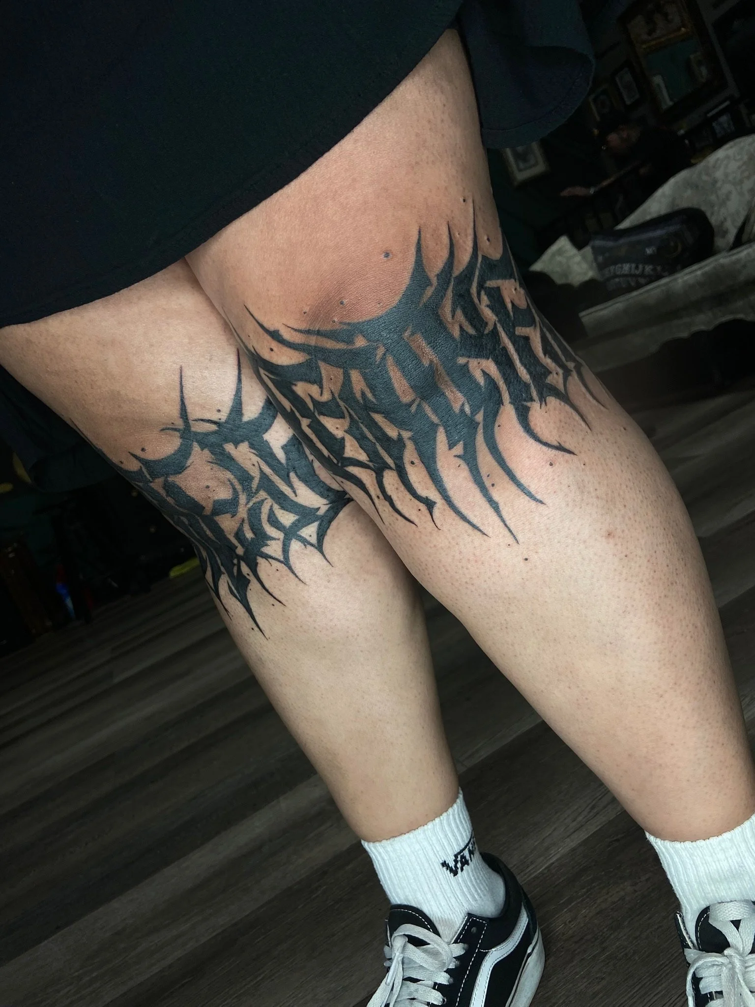 A person's legs wearing black shorts, white socks with black text, and black and white sneakers. The person has a large black tattoo on their thigh.