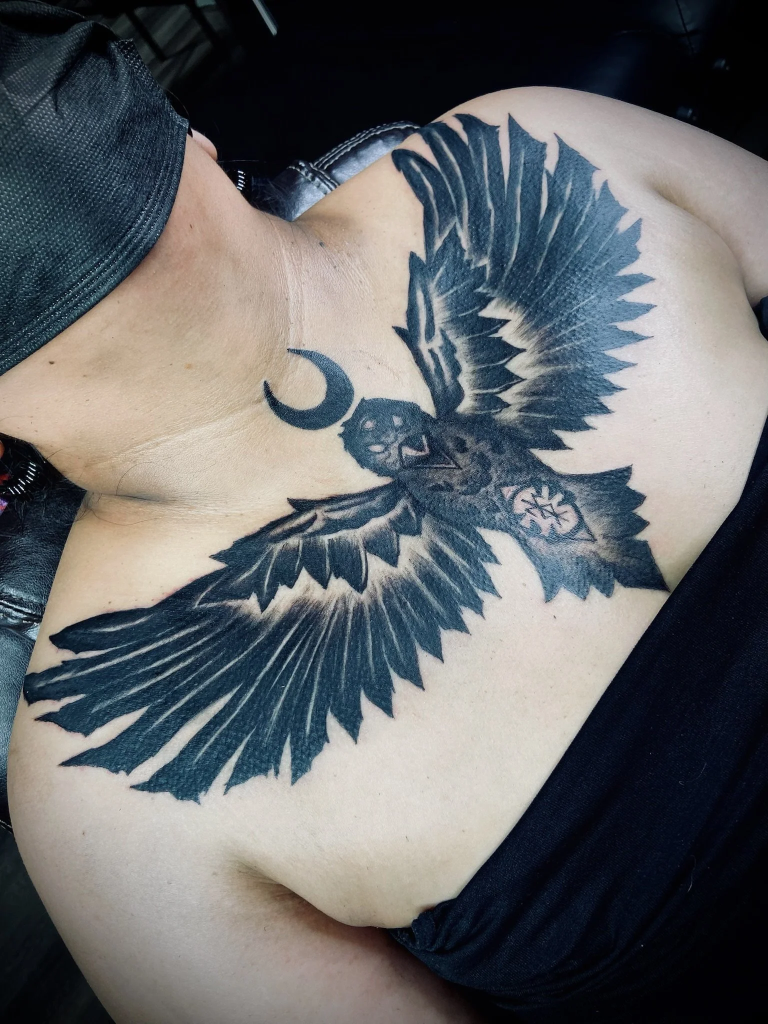 Close-up of a person's side and lower back showing a large black tattoo of an eagle with wings spread, a crescent moon, and a skull with a banner on light skin.