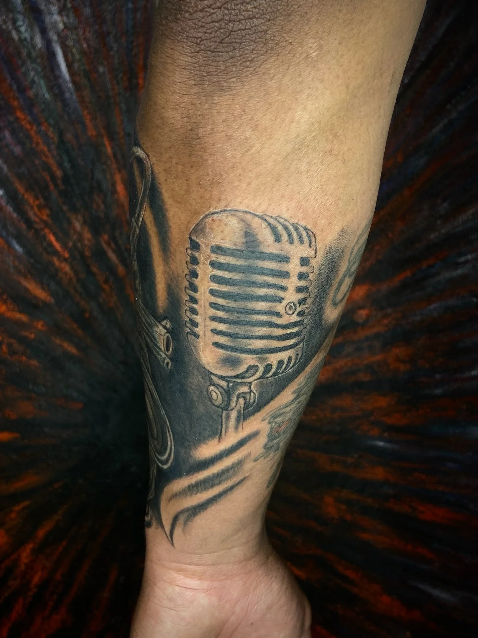 Close-up of a person's lower leg with a tattoo of a vintage microphone on the side, against a dark, striped background.