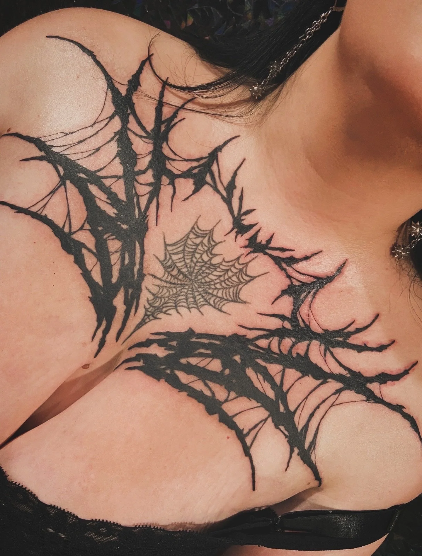 Close-up of a person's shoulder and chest with a black spiderweb tattoo covering the shoulder and extending onto the chest, with detailed web design and black ink.