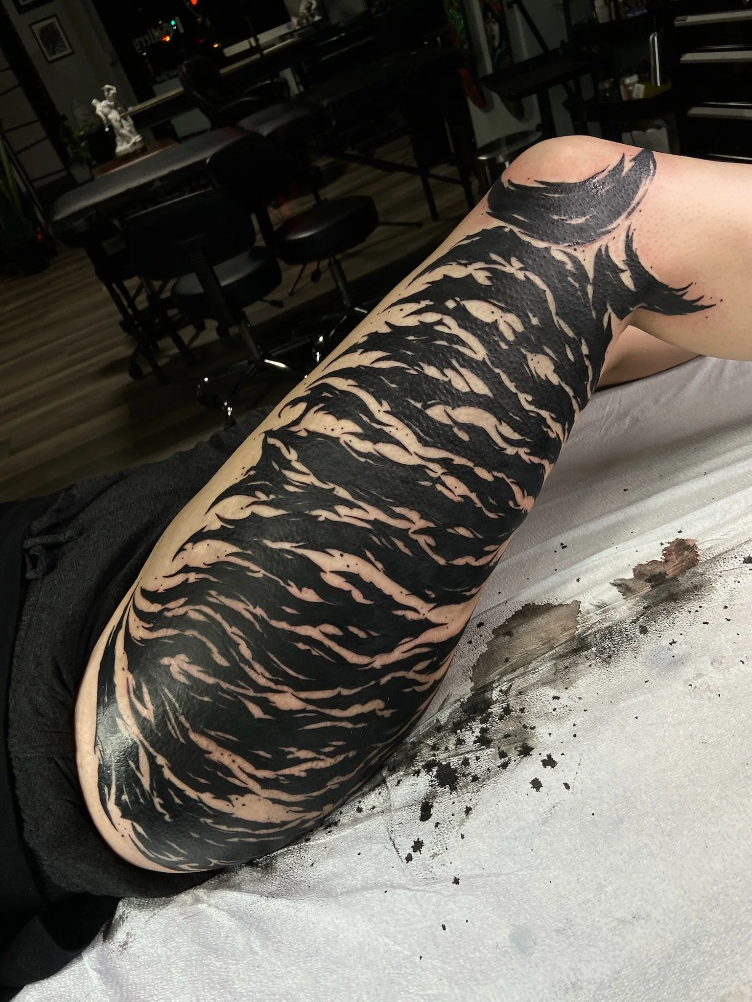 Close-up of a person's arm with a large black abstract tattoo resembling flowing, fiery shapes, extending from the shoulder to the forearm, in a dimly lit room with chairs and tables in the background.