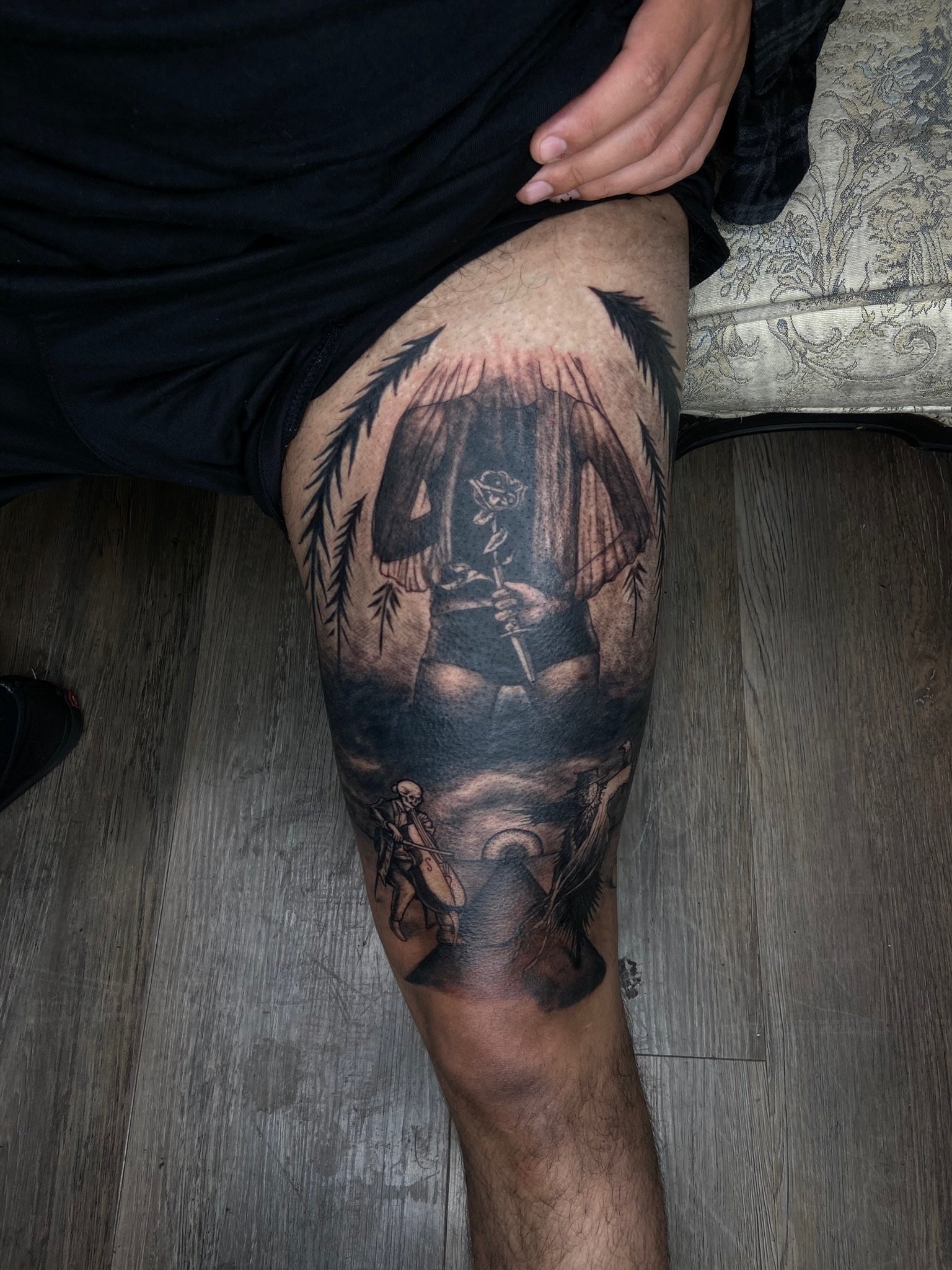 A person's thigh with a large black and gray tattoo of a woman with a bow and arrow, surrounded by feathers and other decorative elements, with the person wearing black shorts and a black shirt, sitting on a wooden floor next to a patterned fabric su