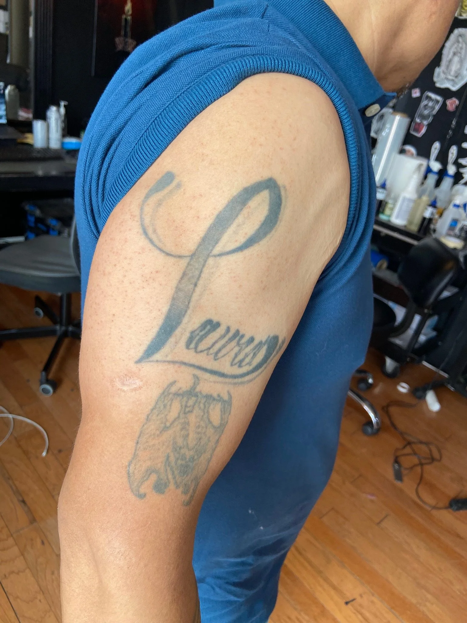 A close-up of a person's upper arm showing a tattoo with cursive writing and a human skull illustration, wearing a blue sleeveless shirt in a room with tattoo supplies and equipment.